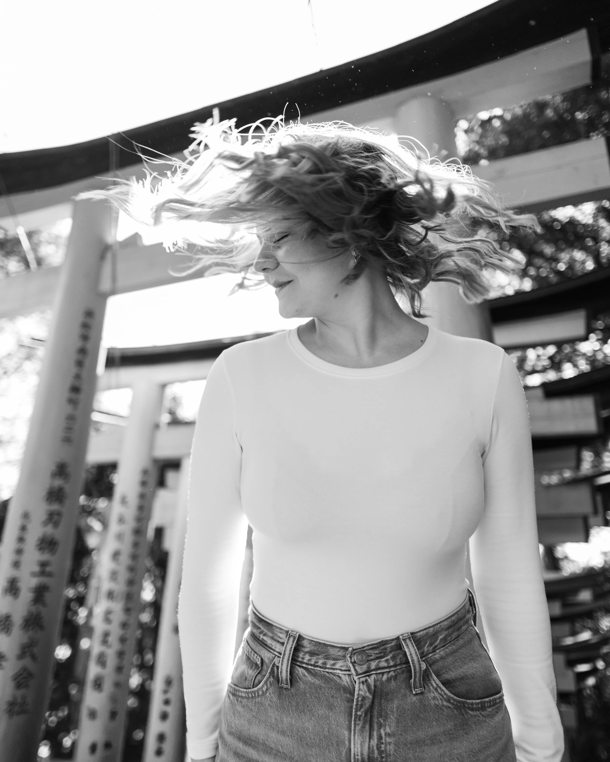 Schwarz-weiße Aufnahme von DominoKati mit lockigem Haar, die ihre Haare im Wind wirbeln lässt. Sie trägt ein weißes longleeves Oberteil und eine Jeans. Im Hintergrund sind Bäume und Tore eines japanischen Tempels in Kyoto erkennbar.