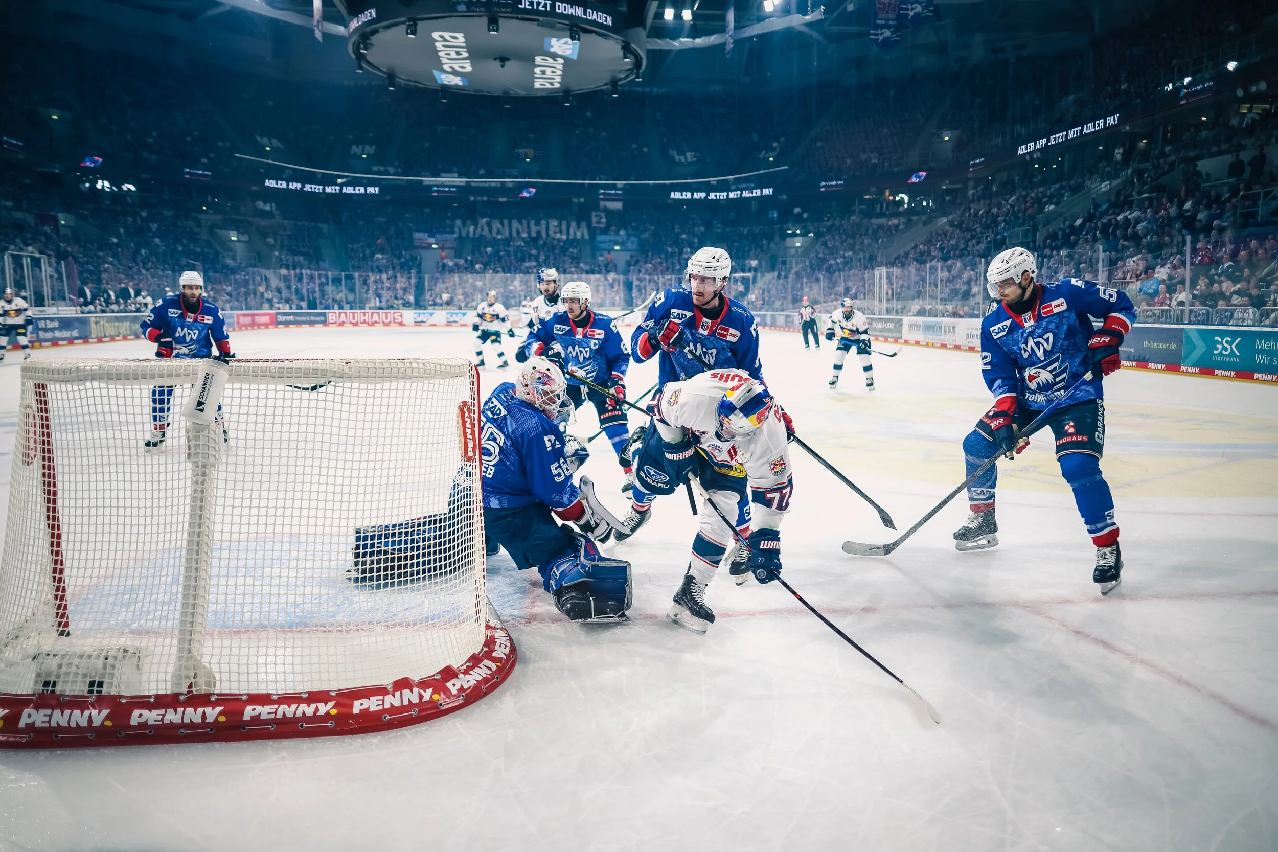 Eishockeyspieler der Adler Mannheim in Aktion vor dem Tor während eines Spiels gegen Red Bull München, mit mehreren Spielern und einem Publikum im Hintergrund. Sportfotografie.