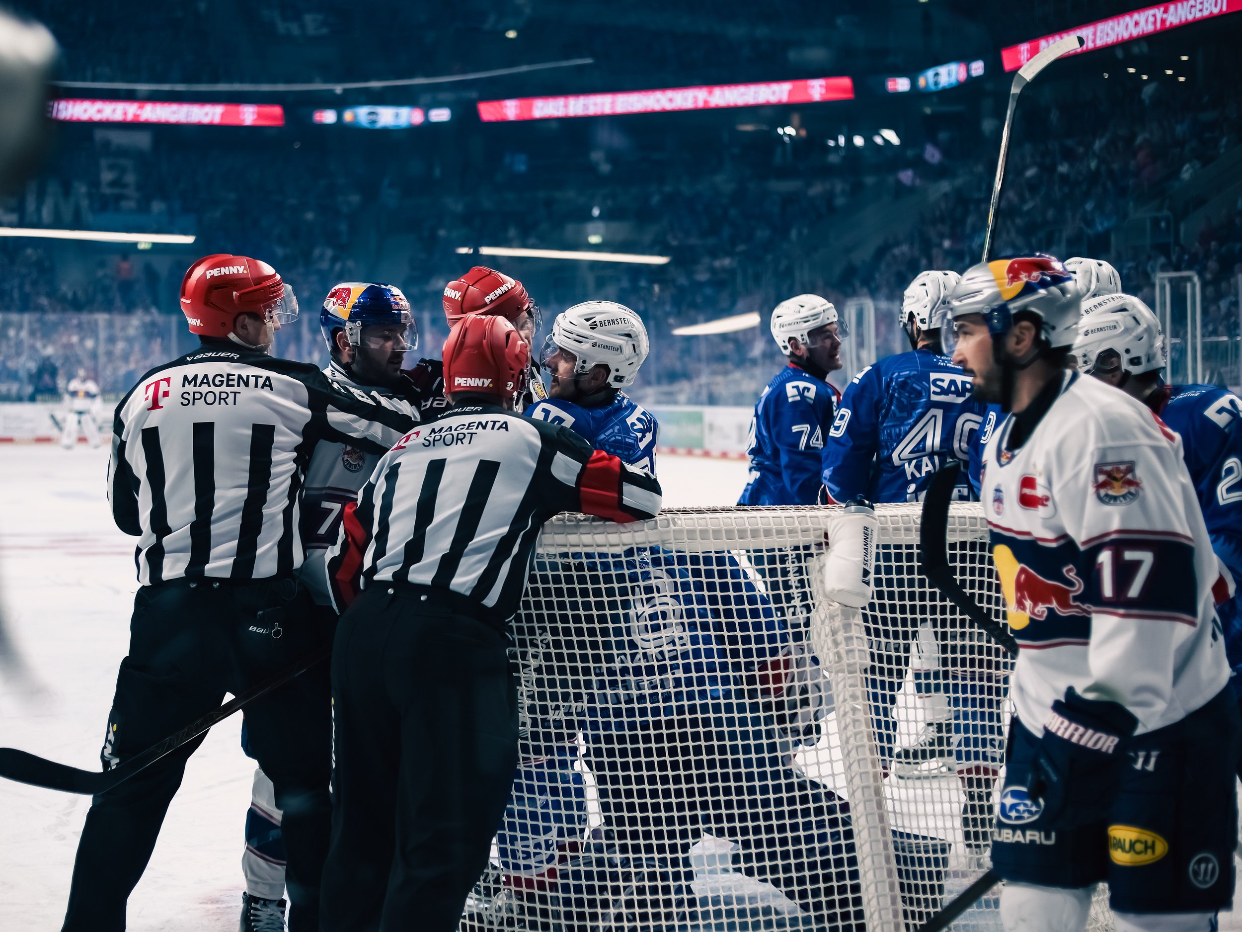 Eishockeyspieler der Adler Mannheim und Red Bull München und Schiedsrichter im Gespräch direkt vor dem Tor auf dem Eis in einem Stadion mit Zuschauern im Hintergrund. Emotionen bei den Spielern. Sportfotografie in Mannheim.