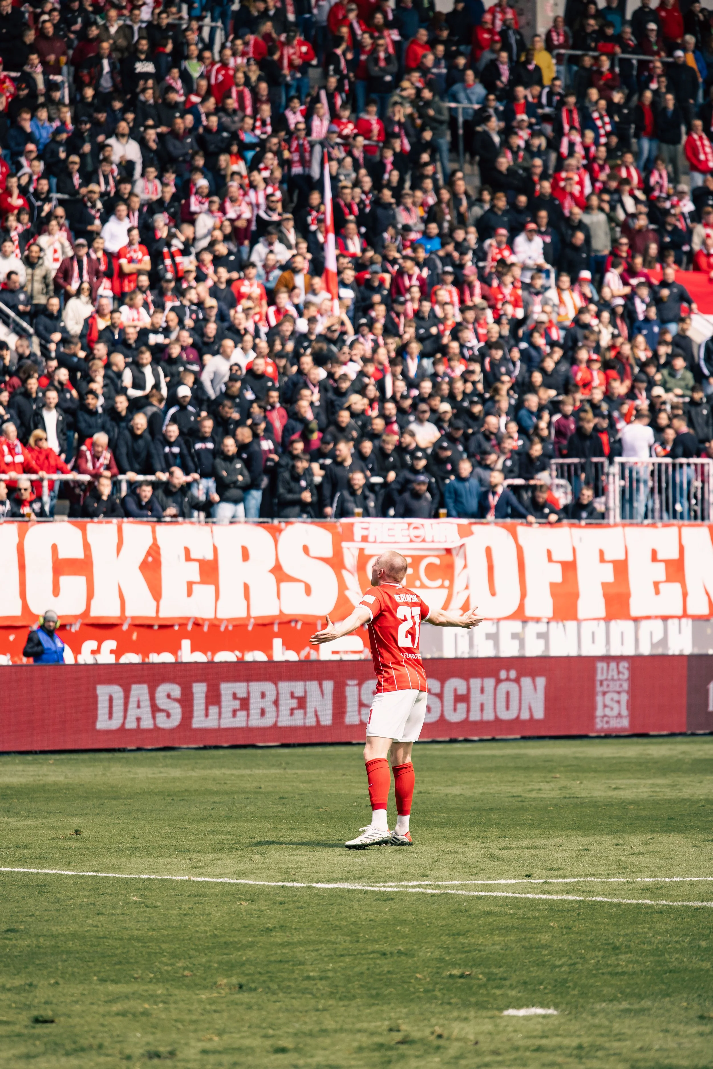 Fußballspieler der Kickers Offenbach in roter Trikotnummer 27 steht auf dem Fußballfeld im Stadion am Biederer Berg, während die Zuschauermenge im Hintergrund jubelt. Sportfotografie bei den Kickers Offenbach, OFC, in der Regionalliga Süd-West.