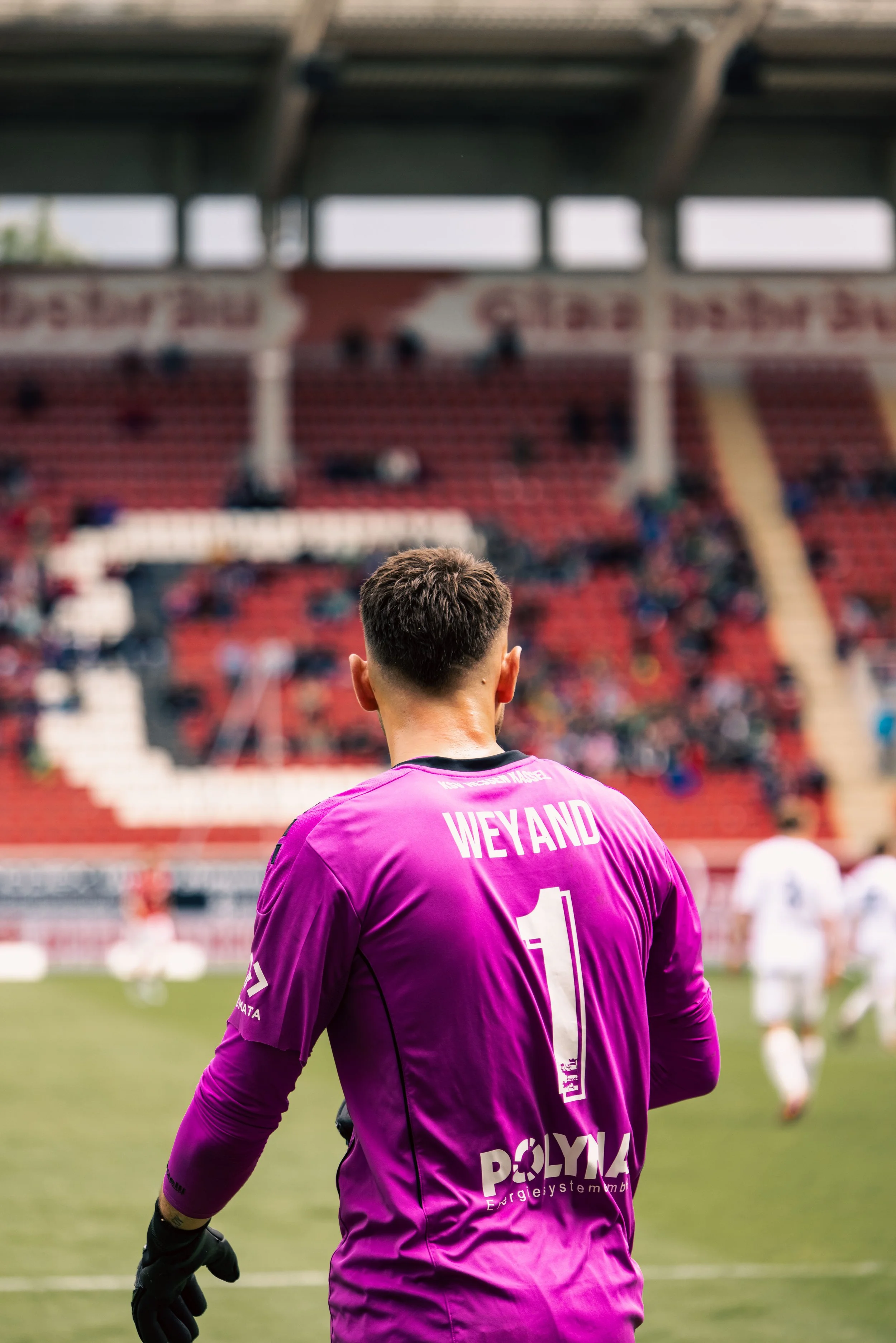 Fußball-Torwart in lila Trikot auf einem Spielfeld mit Zuschauern im Hintergrund.