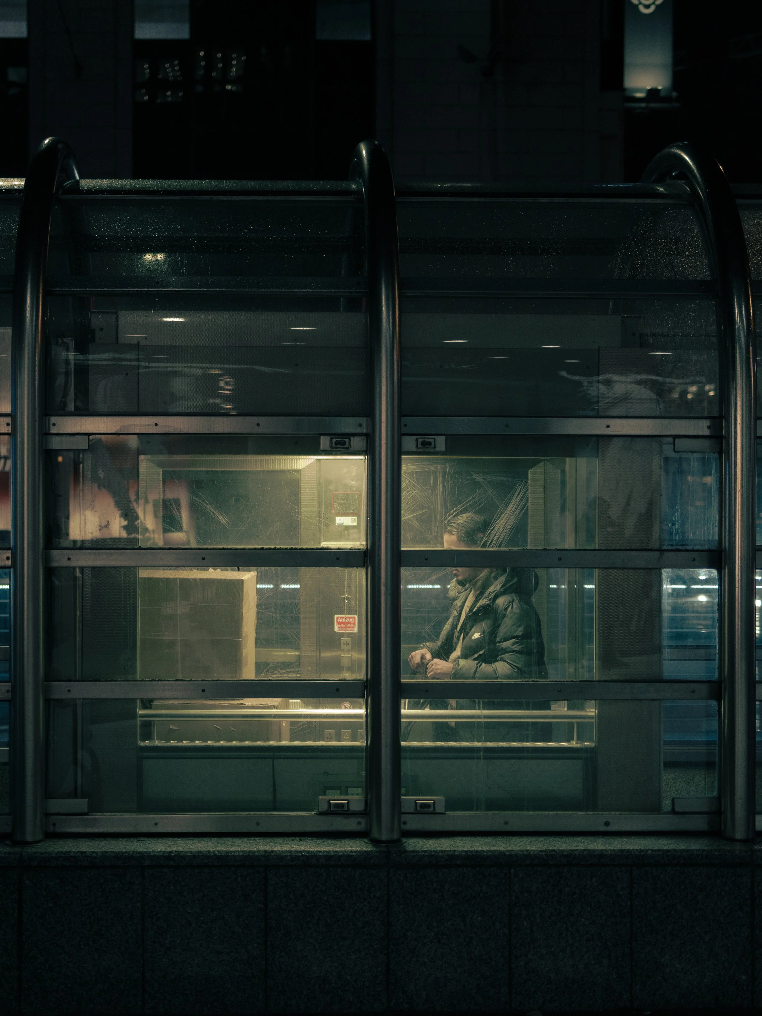 Eine Person in einem Mantel steht in einer Bushaltestelle bei Nacht, die Scheiben sind verschmutzt, im Hintergrund sind Lichter einer Stadt sichtbar. Streetphotography in Düsseldorf. 