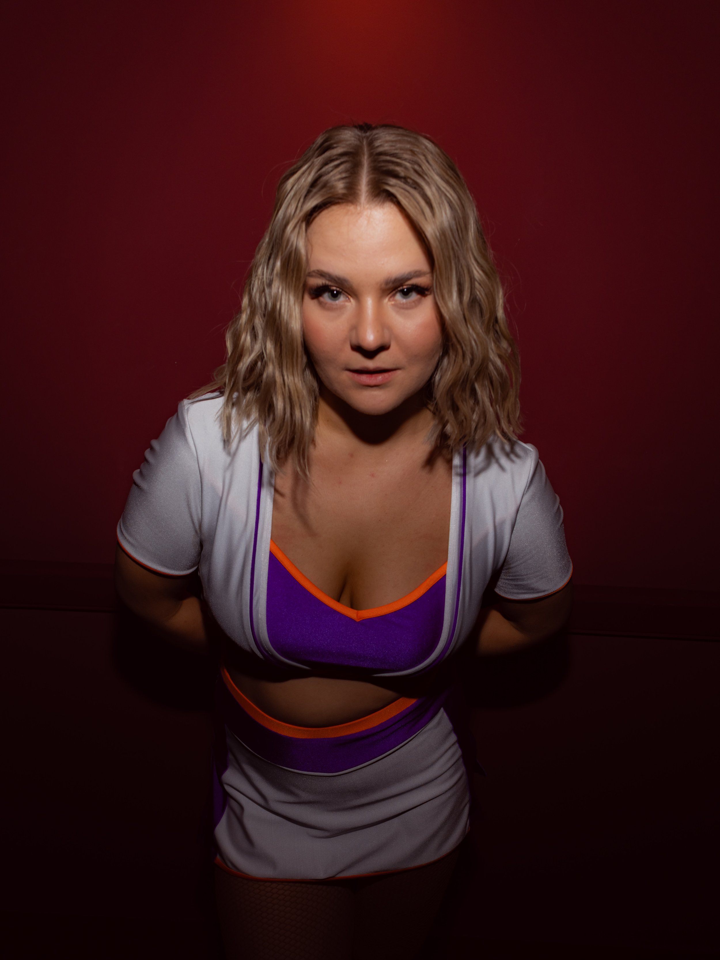 Junge Frau, Cheerleaderin, in Sportkleidung blickt direkt in die Kamera, vor dunklem rotem Hintergrund. Fotografiert mit Blitz.