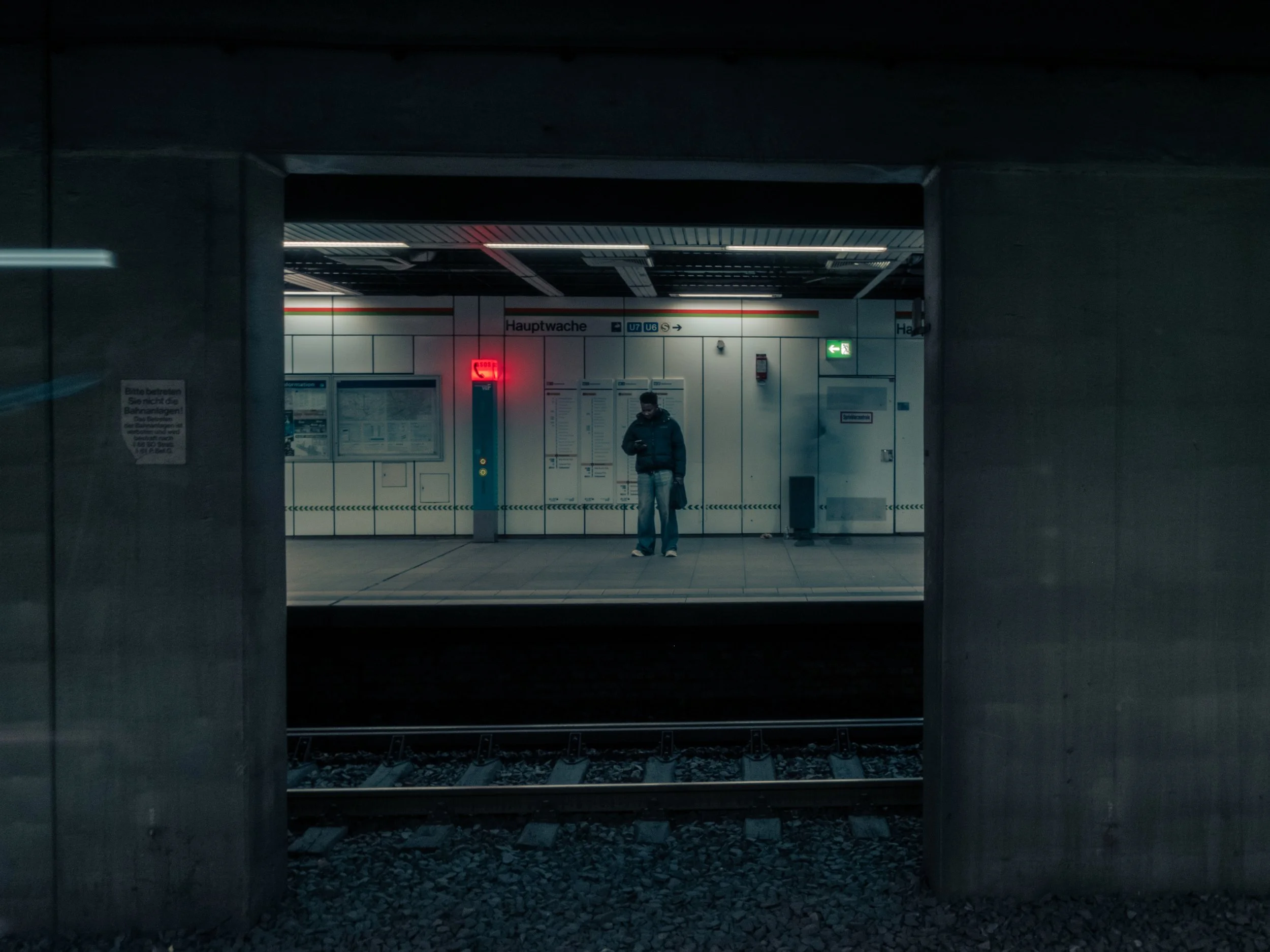 Ein dunkler Bahnhof im Untergrund mit einem einsamen Mann, der auf sein Handy schaut, eine Notrufsäule und einen Notausgangsschilder mit grünem Pfeil sind sichtbar. Der Eingang zum Bahnsteig ist sichtbar im Hintergrund.