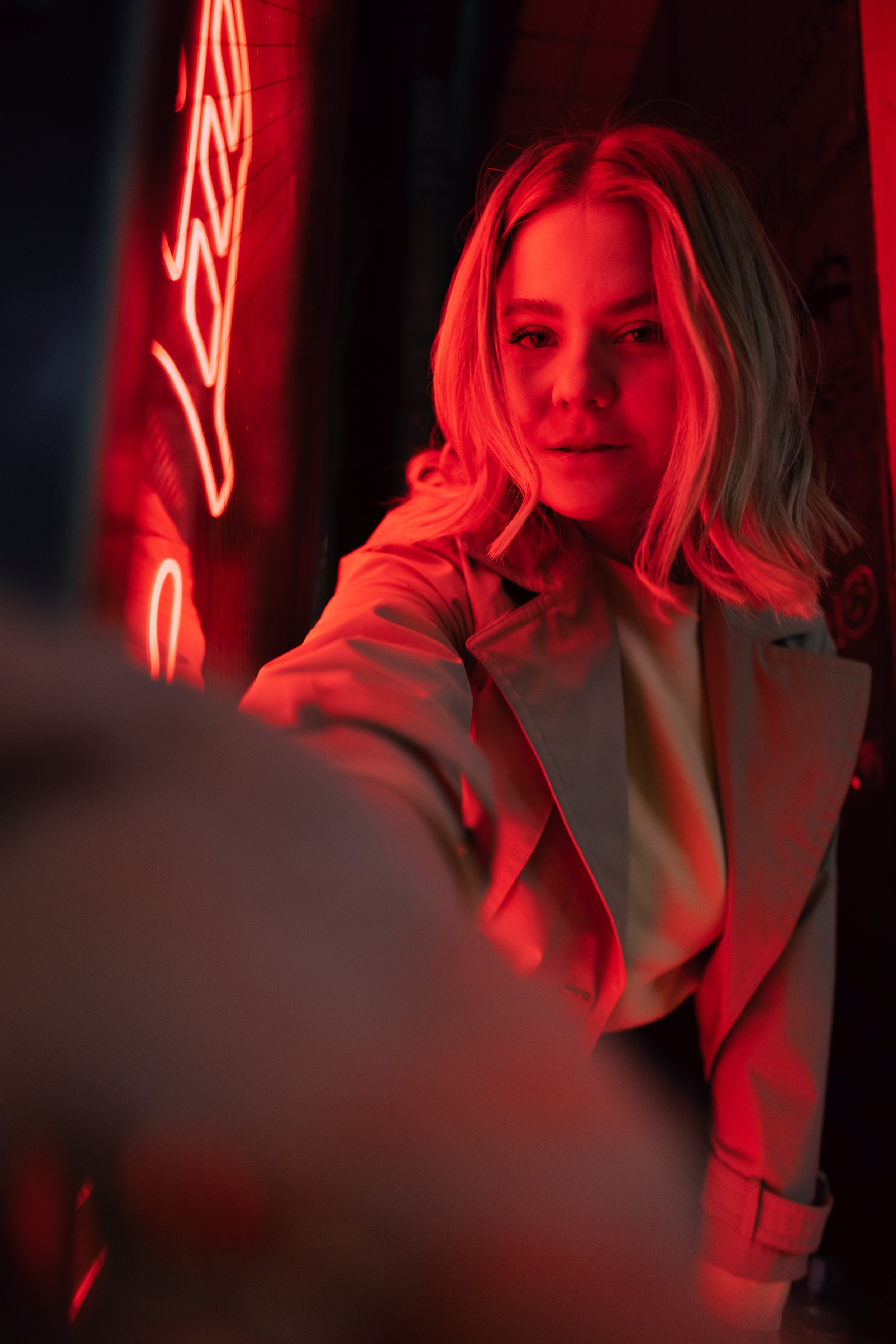 DominoKati schaut in die Kamera, umgeben von rotem Neonlicht. Sie trägt einen Mantel und hat blonde, lockige Haare.