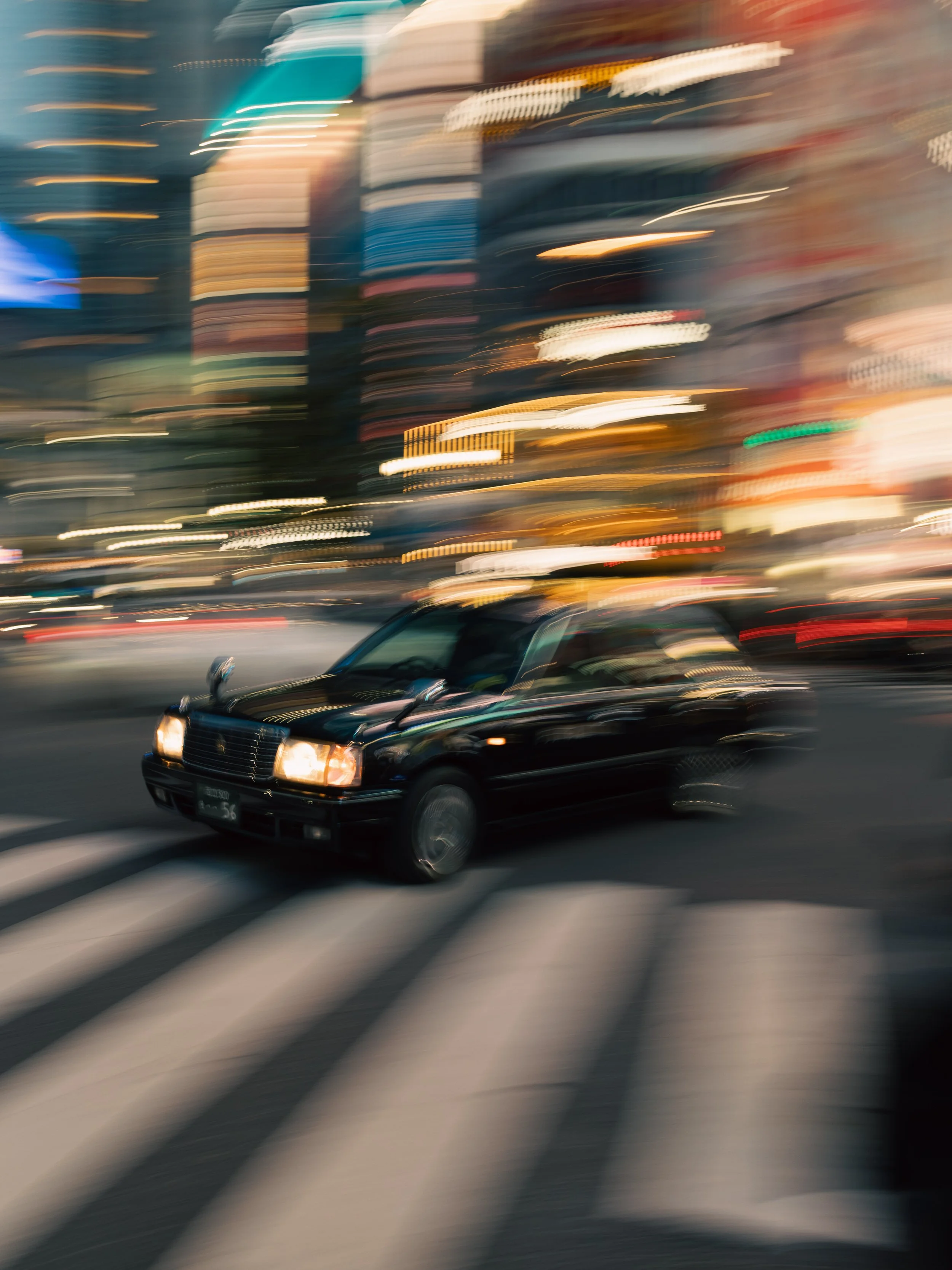 Ein schwarzes Auto fährt bei Nacht durch eine Stadt, während die Lichter von Gebäuden und Straßen im Hintergrund verschwommen sind.