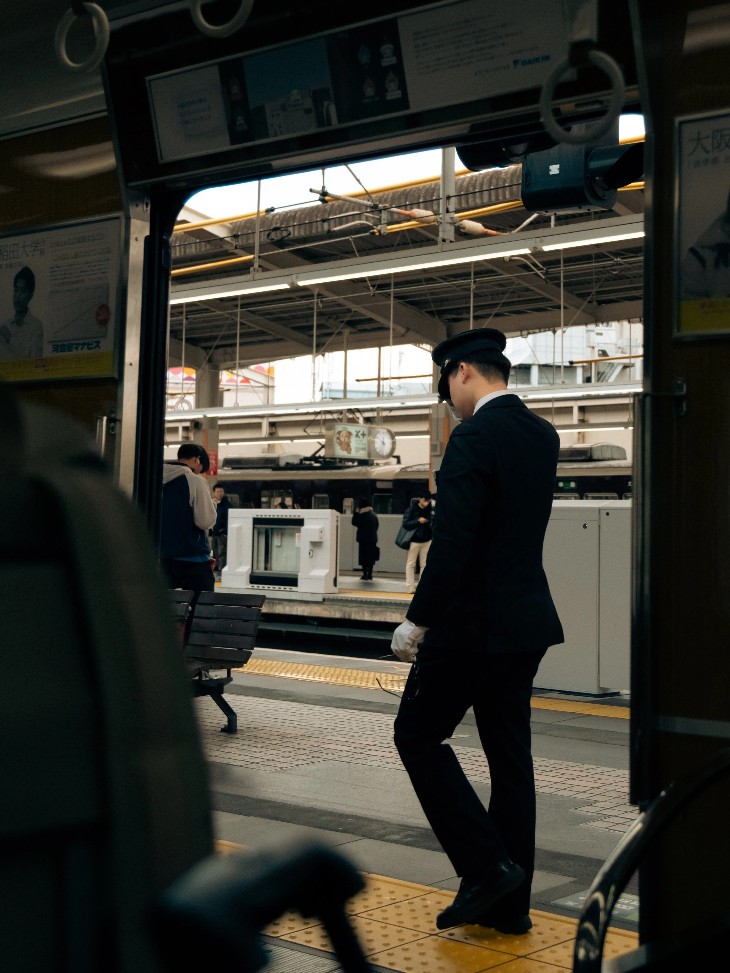 Ein Zugbahnhof in Japan mit einem männlichen Zugbegleiter in Uniform, der auf dem Bahnsteig wartet, fotografiert aus der Sicht eines Bewohners im Zug. Streetphotography.