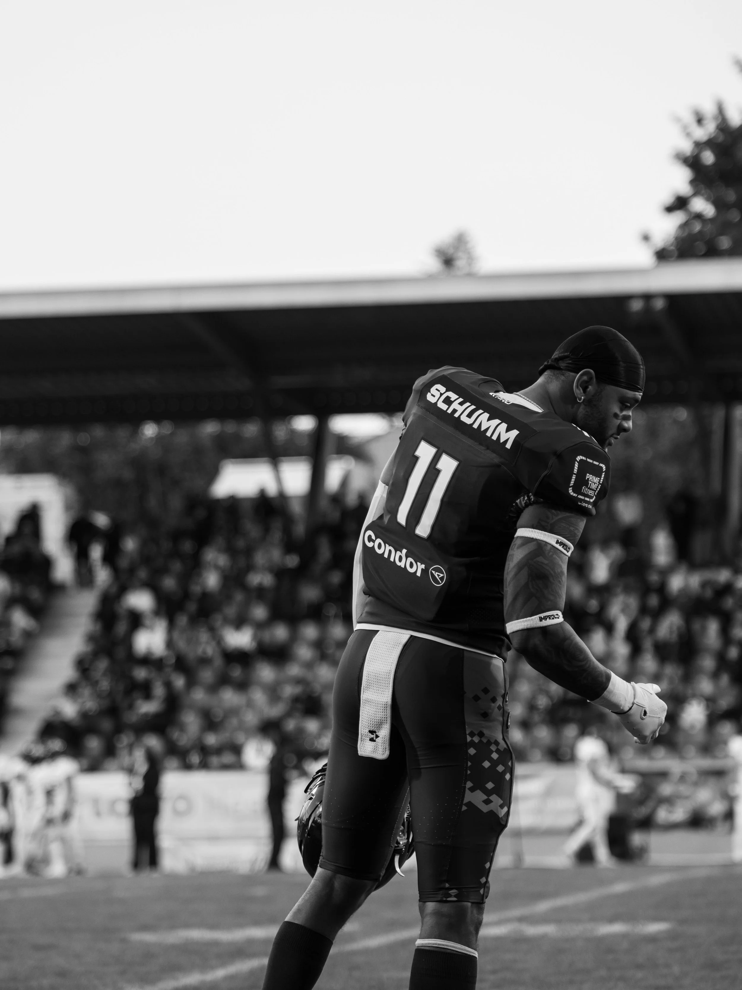 Ein männlicher American-Football-Spieler trägt eine schwarze Uniform mit der Nummer 11 und steht auf dem Spielfeld, im Hintergrund sind Zuschauer und Bänke zu sehen, das Bild ist schwarz-weiß.