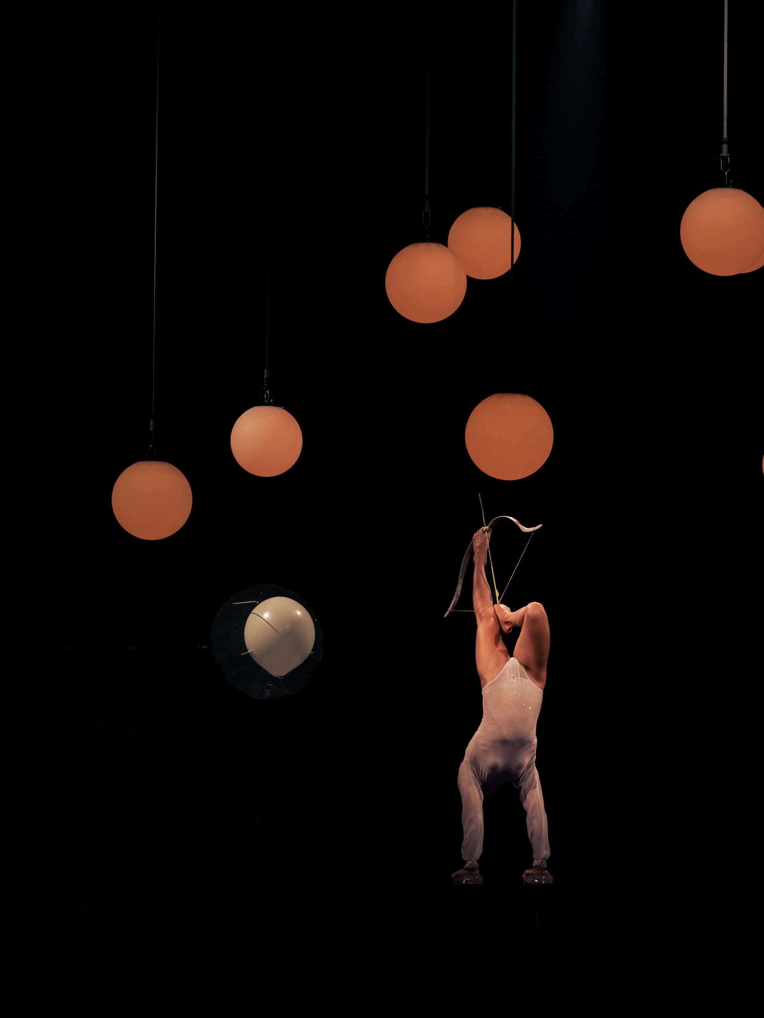 Eine Zirkusartistin schießt Pfeil und Bogen im Handstand mit ihren Füßen, hinter ihr hängen teils orangefarbene, teils weiße und schwarze Lampen vor dunklem Hintergrund, während die Artistin in einem weißen Anzug anhat.