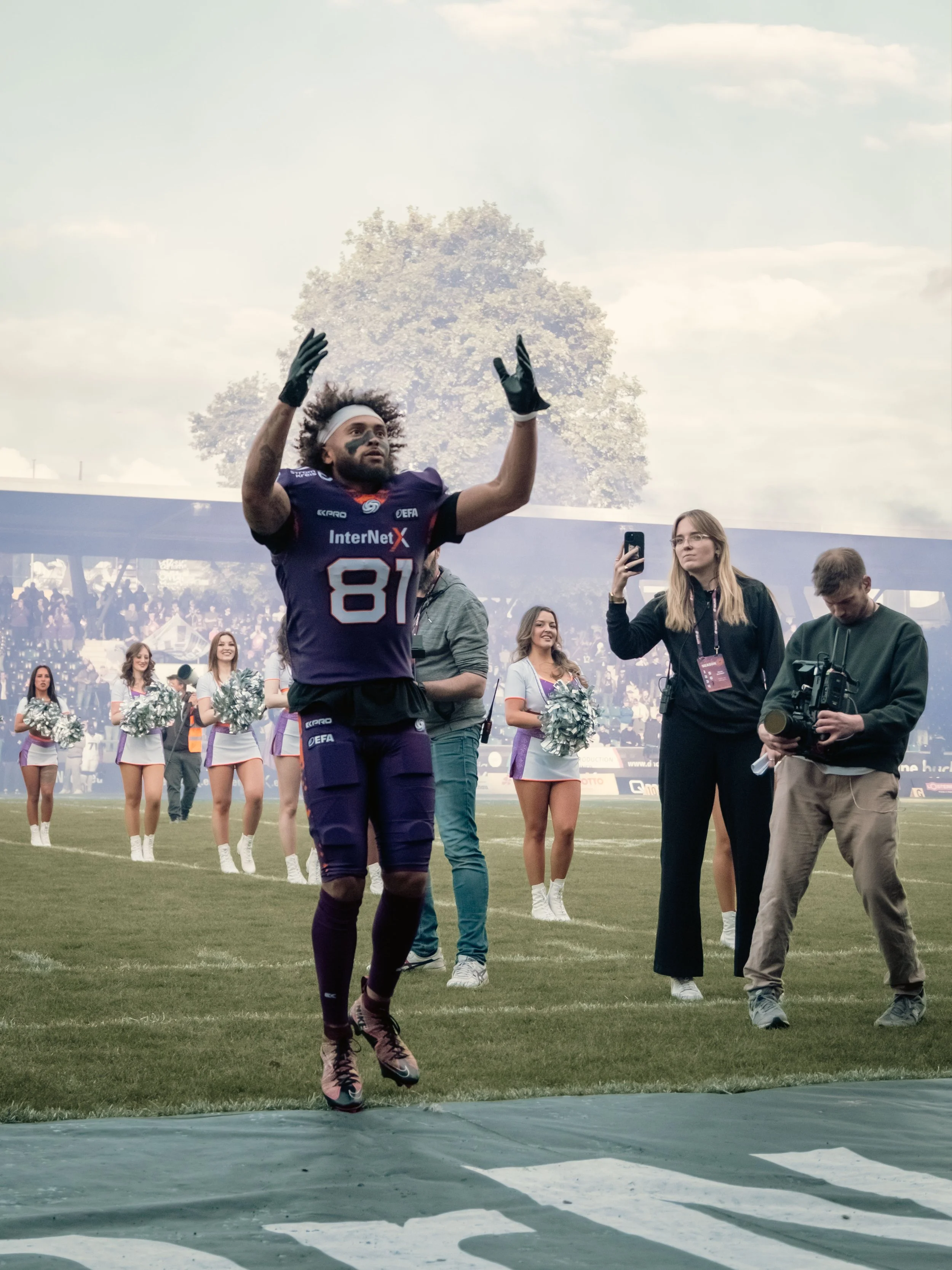 Ein American Football Spieler beim Jubel, während im Hintergrund Cheerleader und andere Personen auf dem Spielfeld stehen, im Stadion mit Zuschauern im Hintergrund. Sportfotografie in Frankfurt.