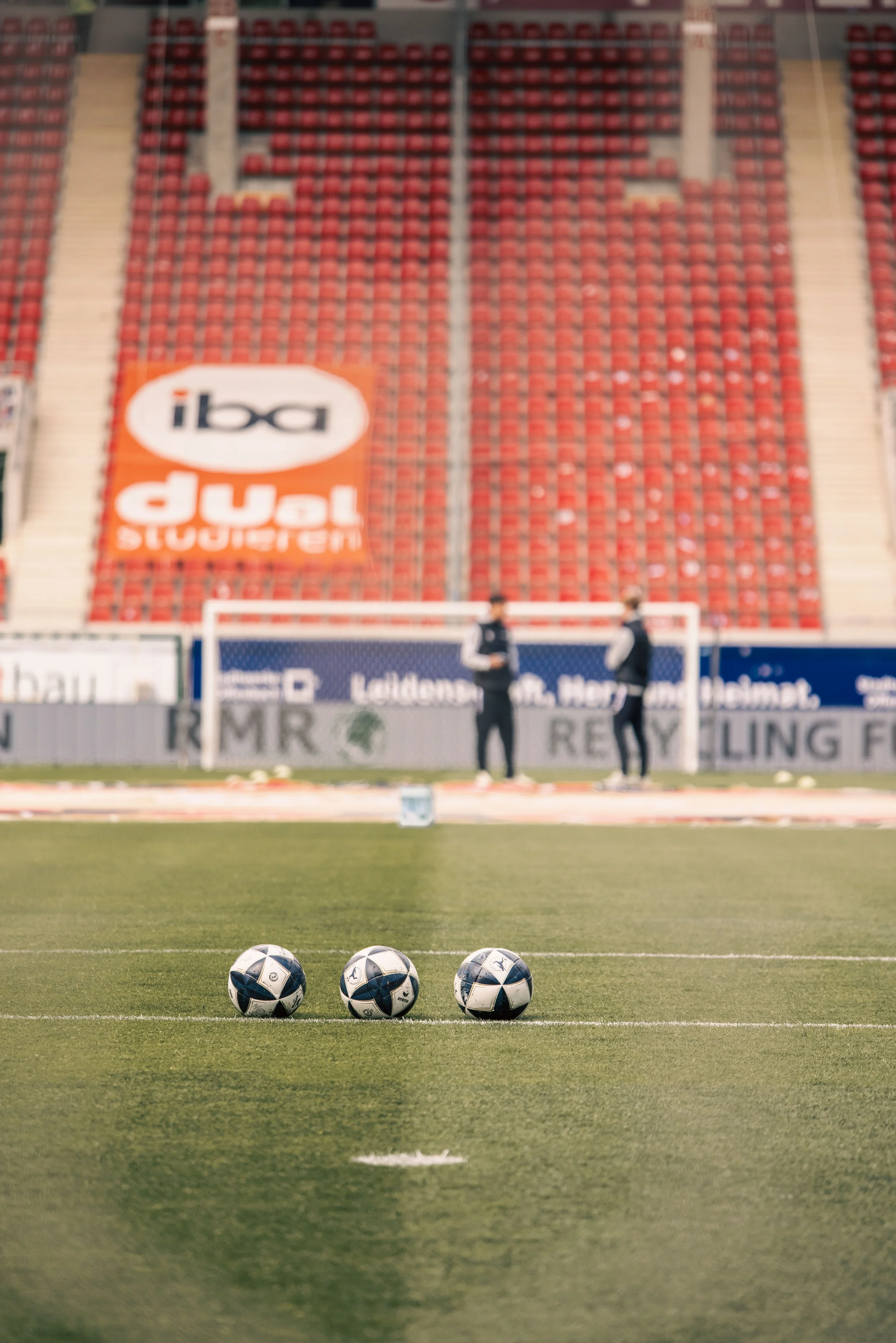 Drei Fußballbälle auf dem Spielfeld vor einer Torlinie, im Hintergrund zwei Personen im Gespräch, leere rote Sitzreihen im Stadion.