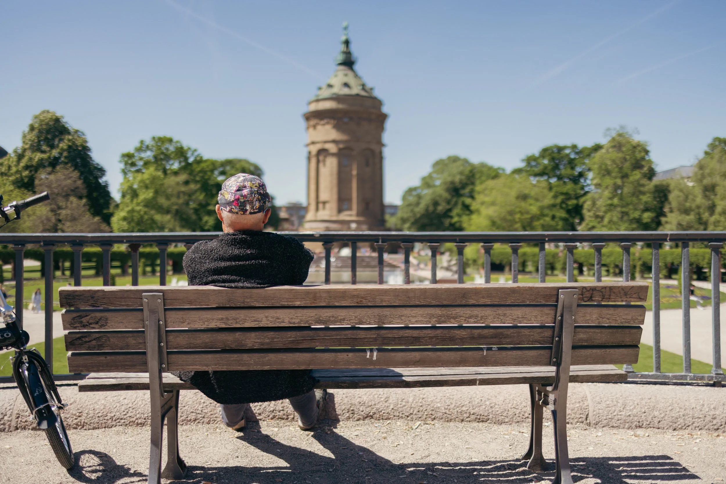 Eine Person mit buntem Hut sitzt auf einer Holzbank und schaut auf den Mannheimer Wasserturm in einem Park, umgeben von Bäumen, an einem sonnigen Tag.