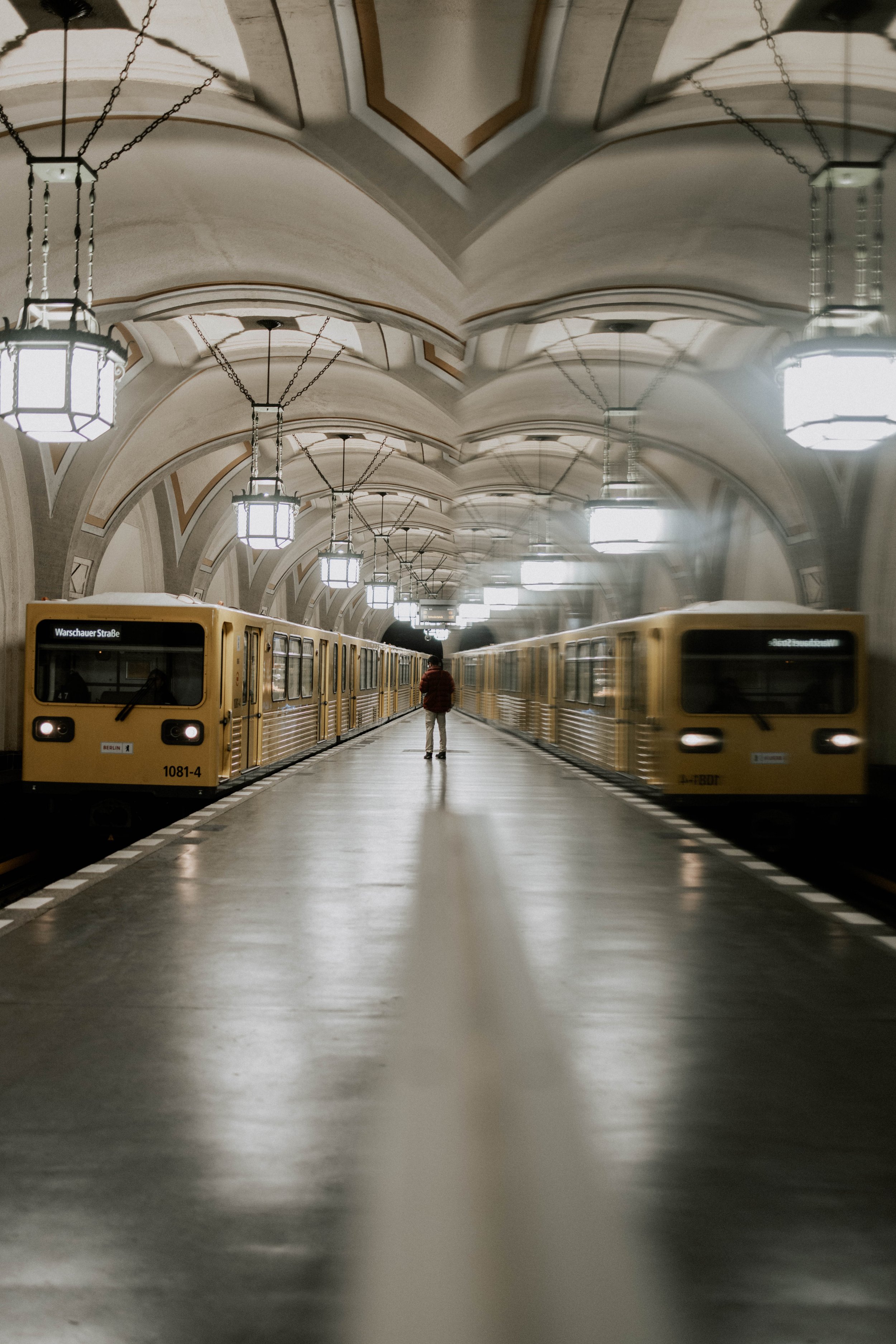 Blick in eine U-Bahn-Station in Berlin mit gelben Zügen, hoher gewölbter Decke mit Kronleuchtern, eine einzelne Person steht auf den Gleisen. Spiegelung fotografiert als Streetphotography in Berlin.