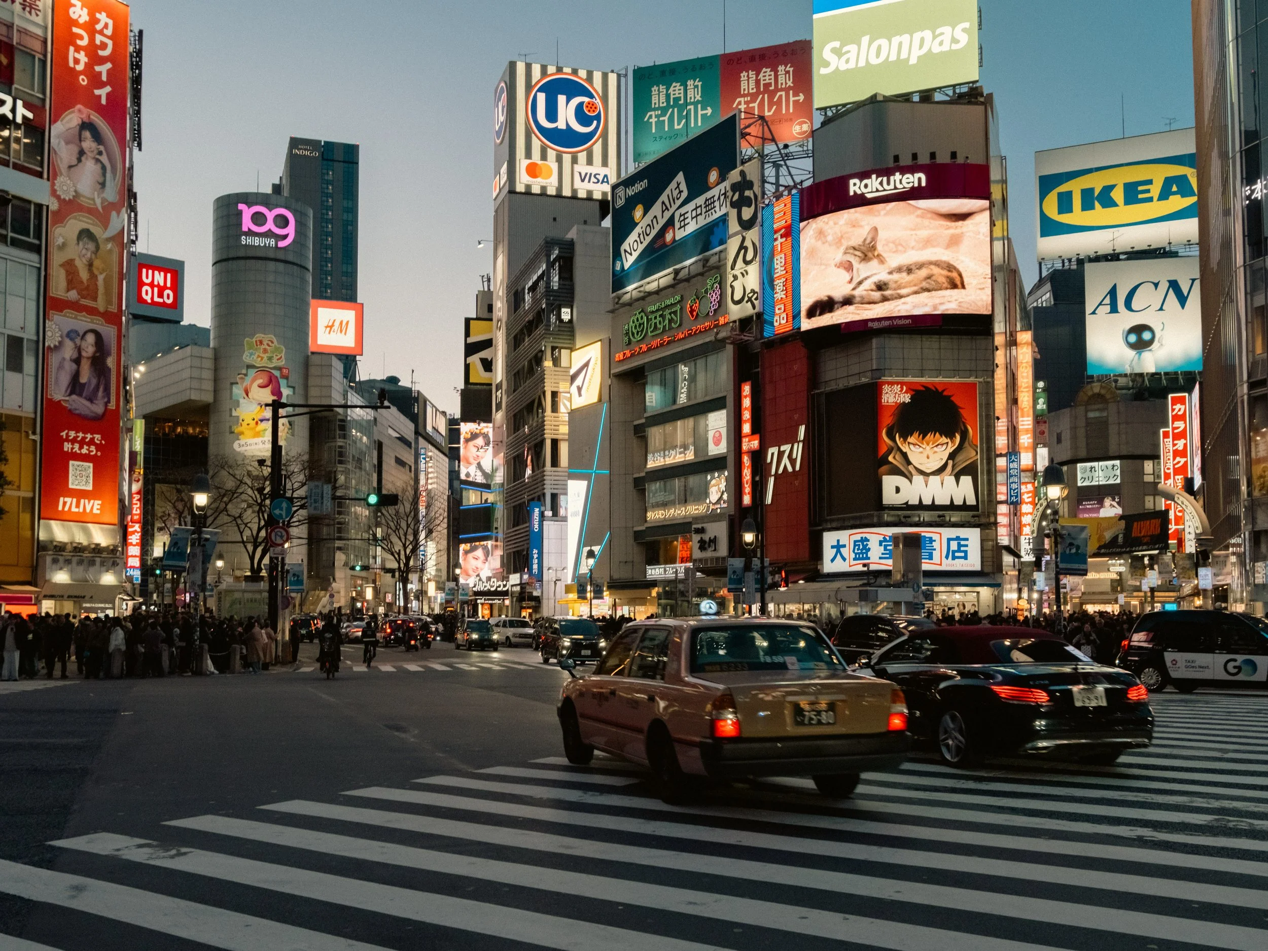 Stadtbild mit vielen beleuchteten Werbetafeln und Hochhäusern bei Dämmerung an der Shibuya Kreuzung in Tokio, Straßenverkehr mit Autos, Menschen überqueren die Straße. Street-Fotografie in Tokio, Japan.