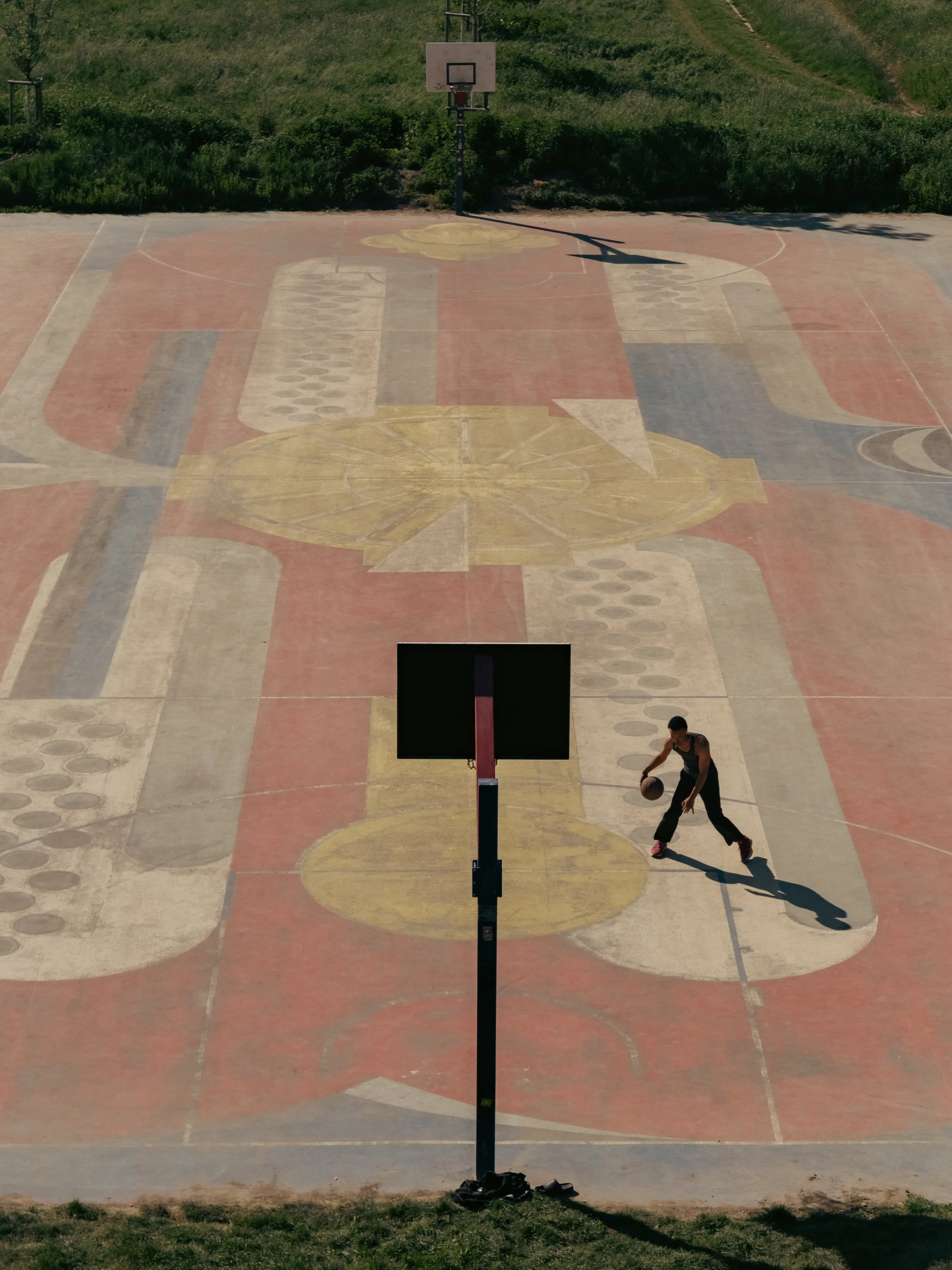 Ein Basketballplatz im Freien mit einem Basketballspieler, der einen Ball dribbelt, während die Sonne Schatten wirft, im Hintergrund grüne Büsche.