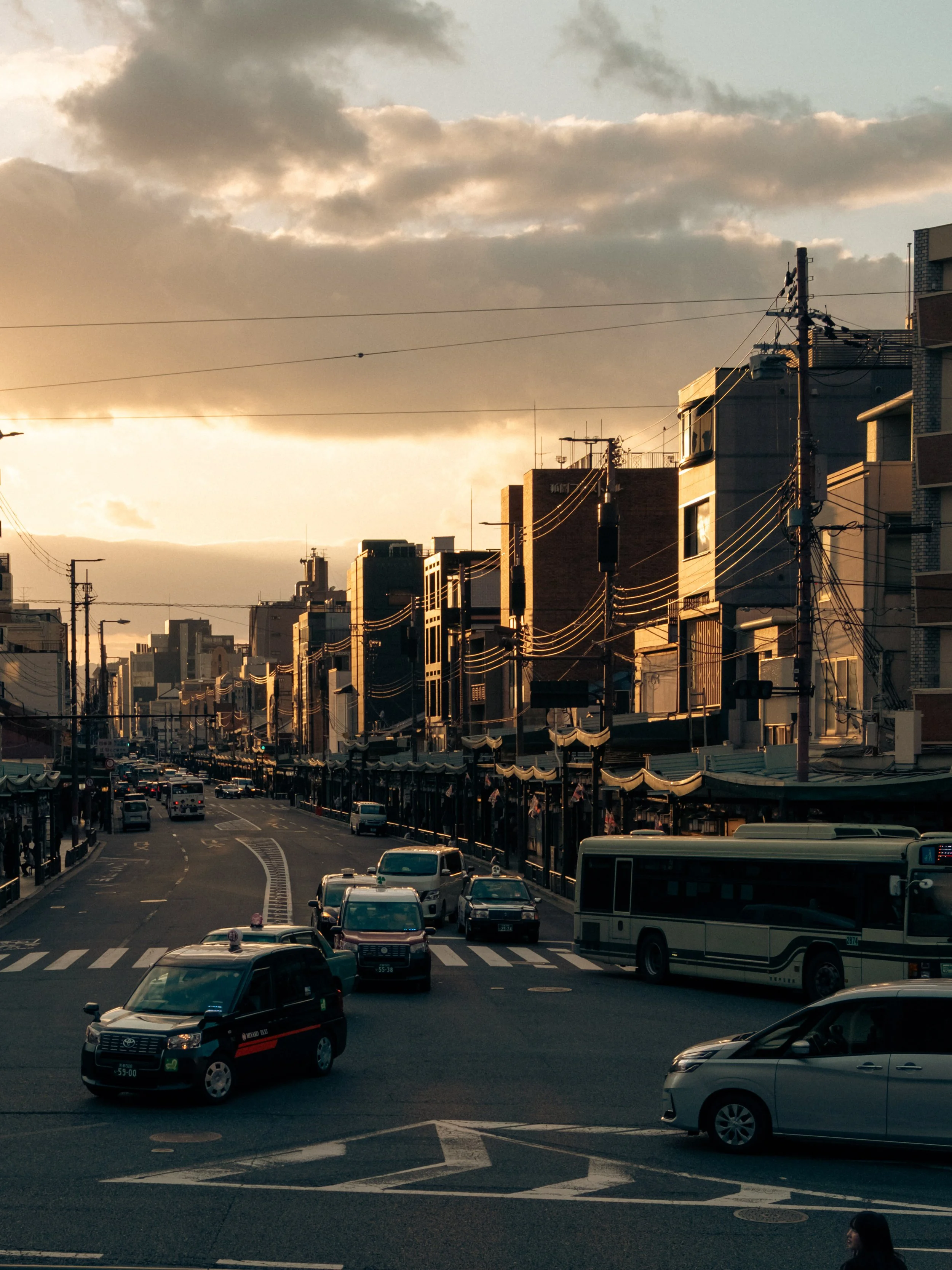Stadtverkehr mit Autos, Bus, Hochhäuser und Stromleitungen bei Sonnenuntergang, Wolken am Himmel.