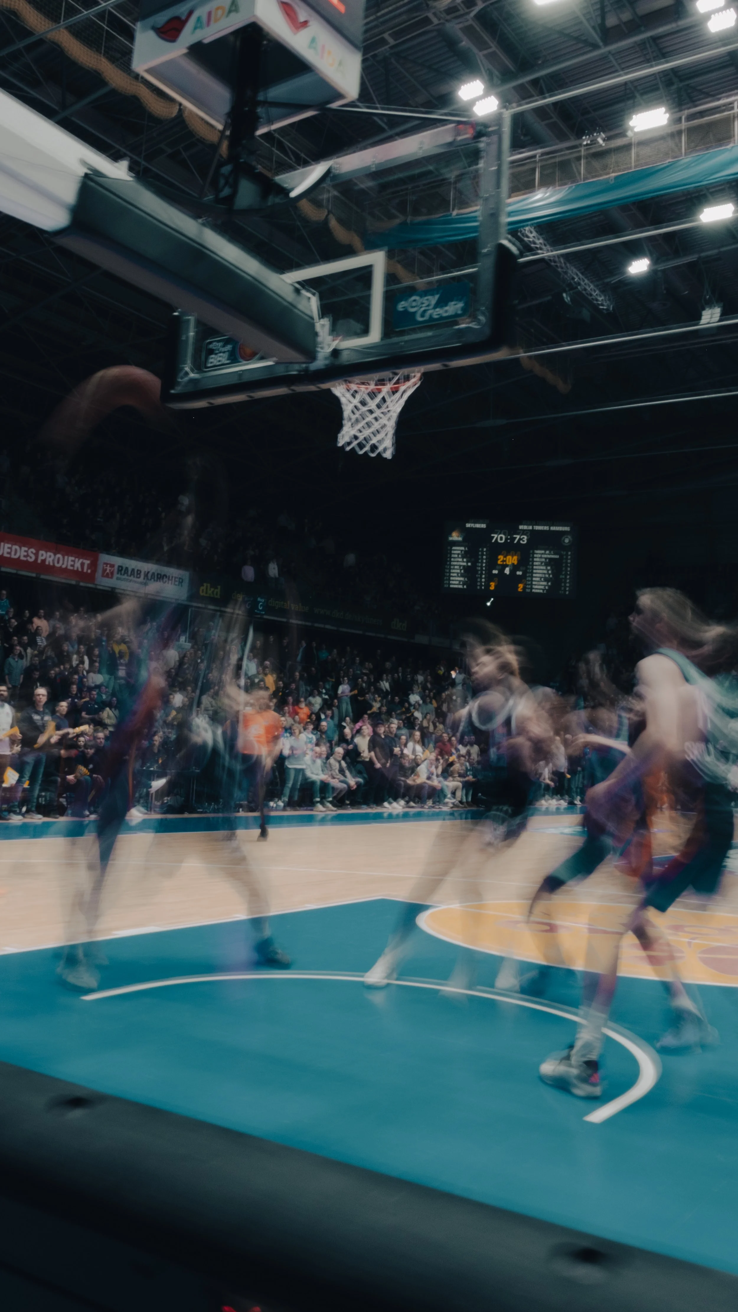 Blick auf ein Basketballspiel in einer Halle, auf dem Korb sind Spieler, das Publikum sitzt im Hintergrund, die Anzeigetafel zeigt Spielstand und Zeit. Aufgenommen mit Langzeitbelichtung bei den Frankfurt Skylines.