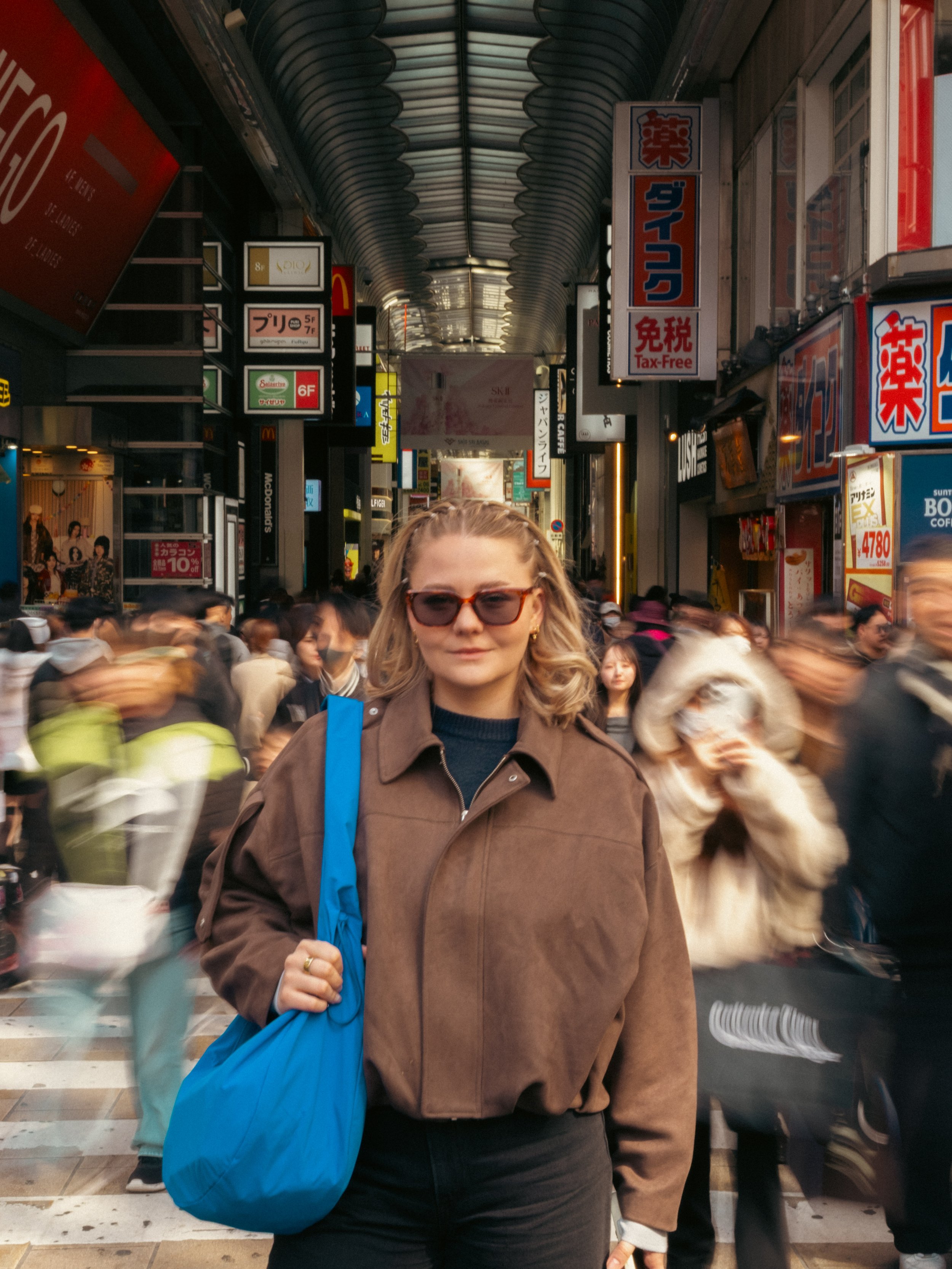 Eine Frau mit Brille und braunkleidung steht in einer belebten Einkaufsstraße in Japan, umgeben von Menschen, Werbetafeln und Geschäften.