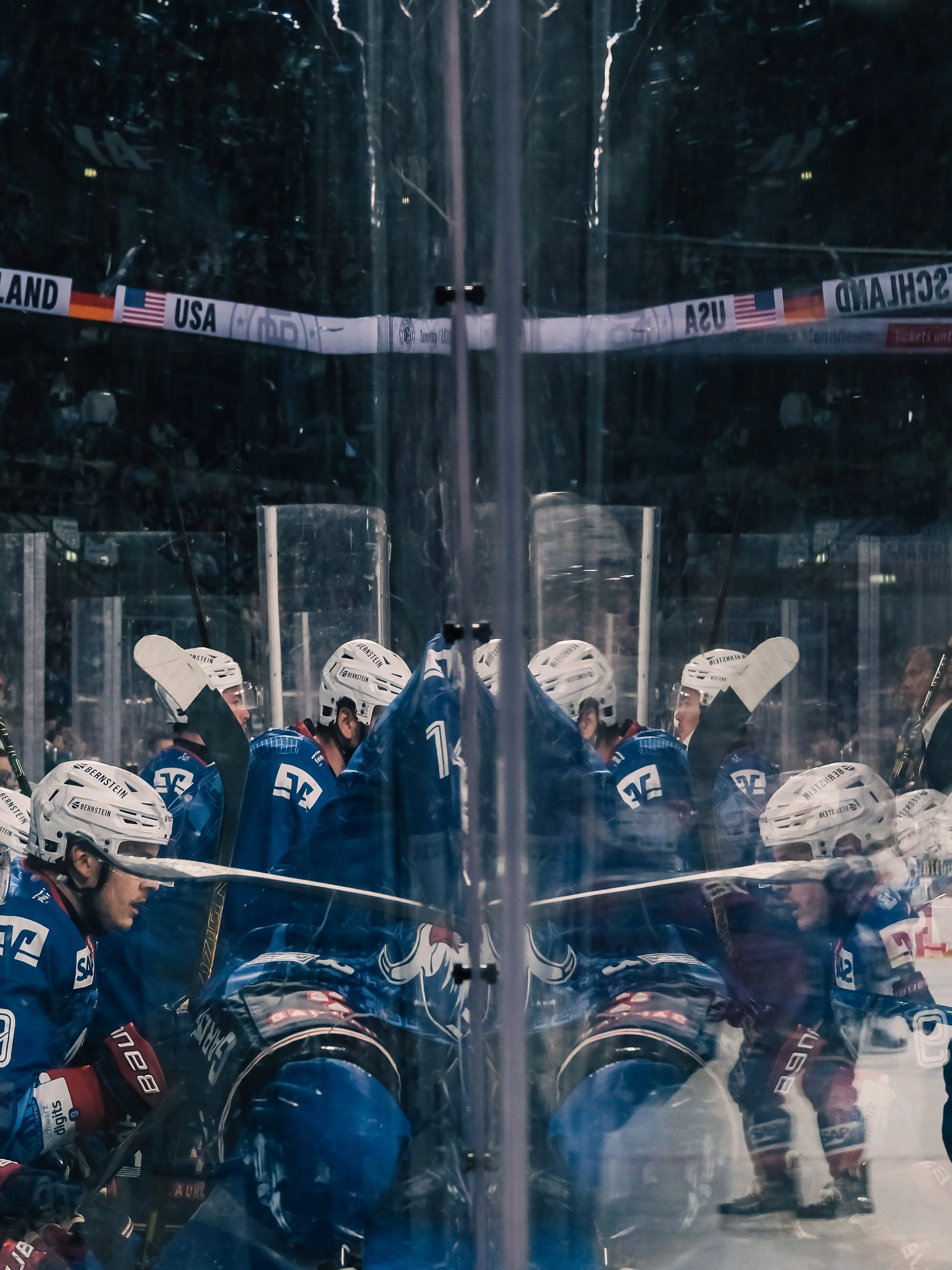 Eishockeyspieler in blauen Trikots auf der Bank während eines Spiels, mit Spielernamen auf Helmen sichtbar. Sportfotografie in Mannheim.
