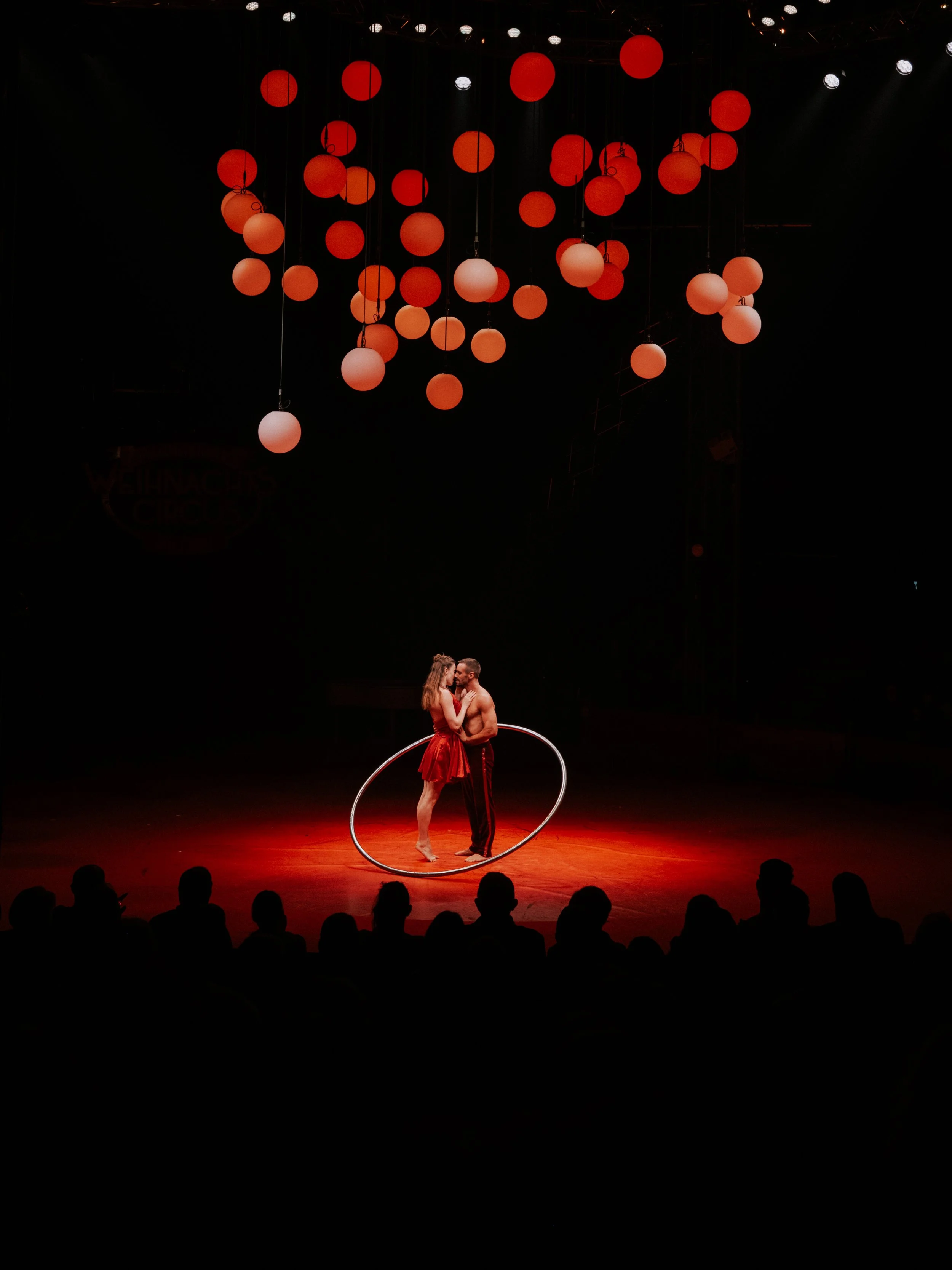 Ein Zirkus-Performance mit einem Paar, das in der Mitte der Bühne steht, umkreist von einem Car-Rad, während über ihnen rote und orangefarbene Lampen hängen; das Publikum schaut zu.