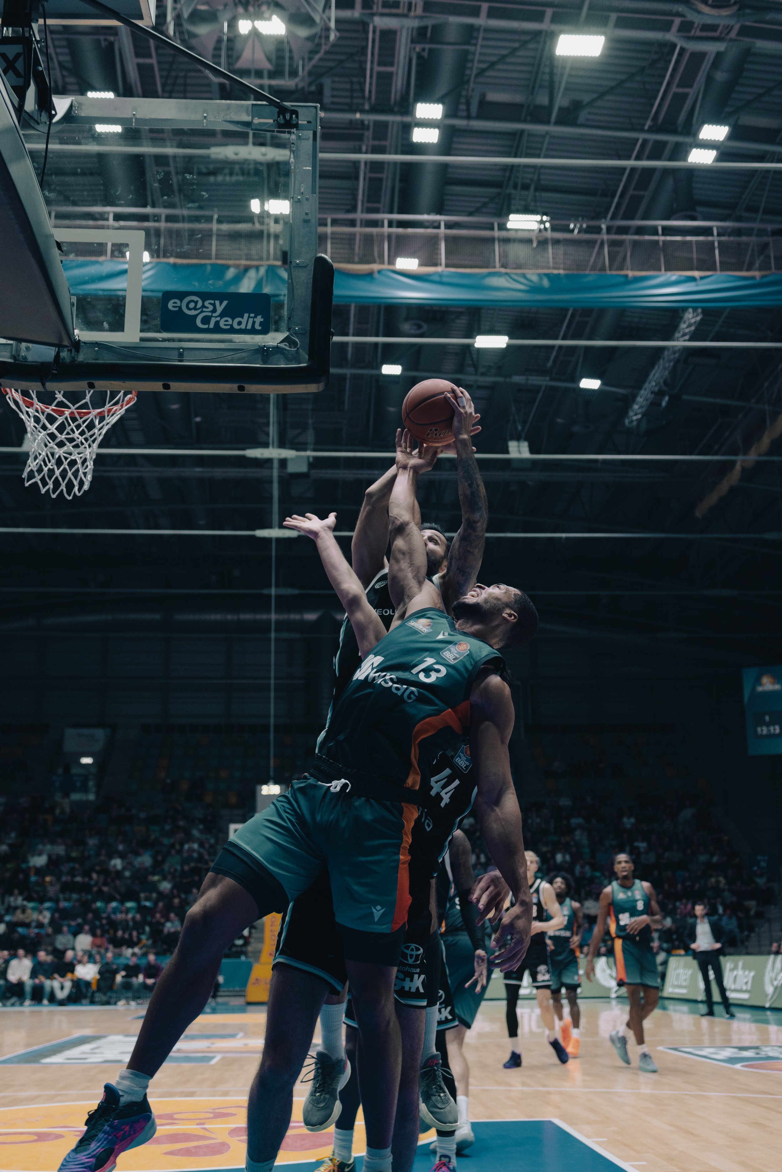 Zwei Basketballspieler der Frankfurt Skyliners kämpfen um den Ball in einem Basketballspiel in einer Sporthalle, mit anderen Spielern und Zuschauern im Hintergrund. Sportfotografie bei den Frankfurt Skyliners.