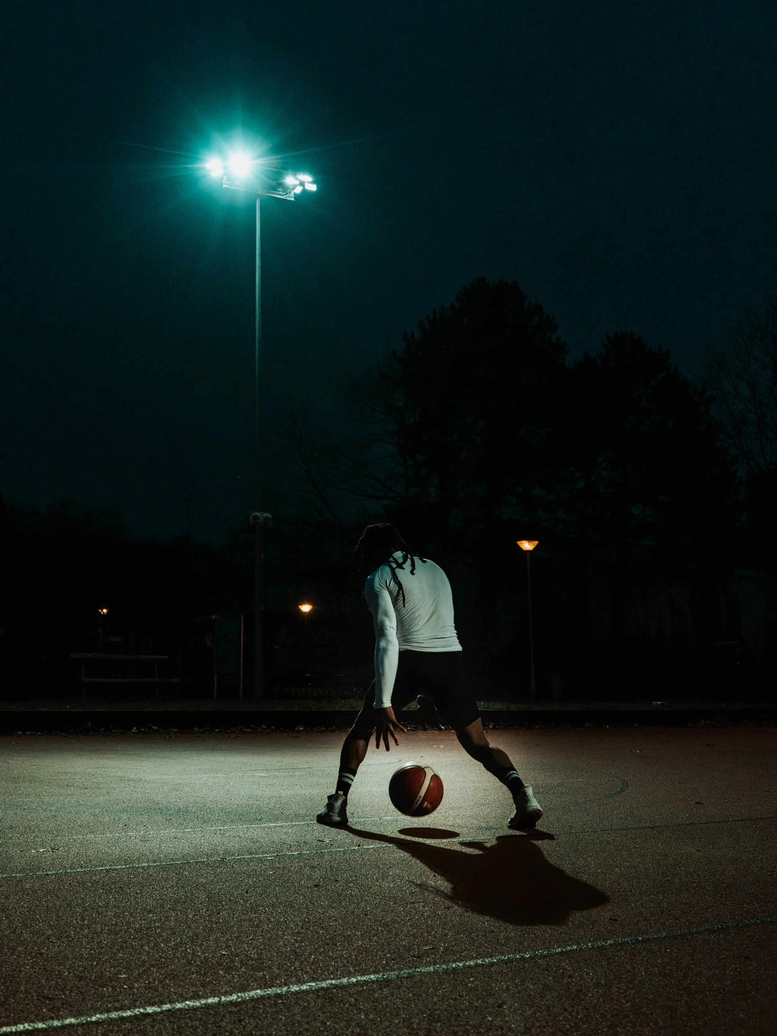 Ein junger Mann spielt Basketball auf einem dunklen Outdoor-Court bei Nacht, beleuchtet von Straßenlaternen.
