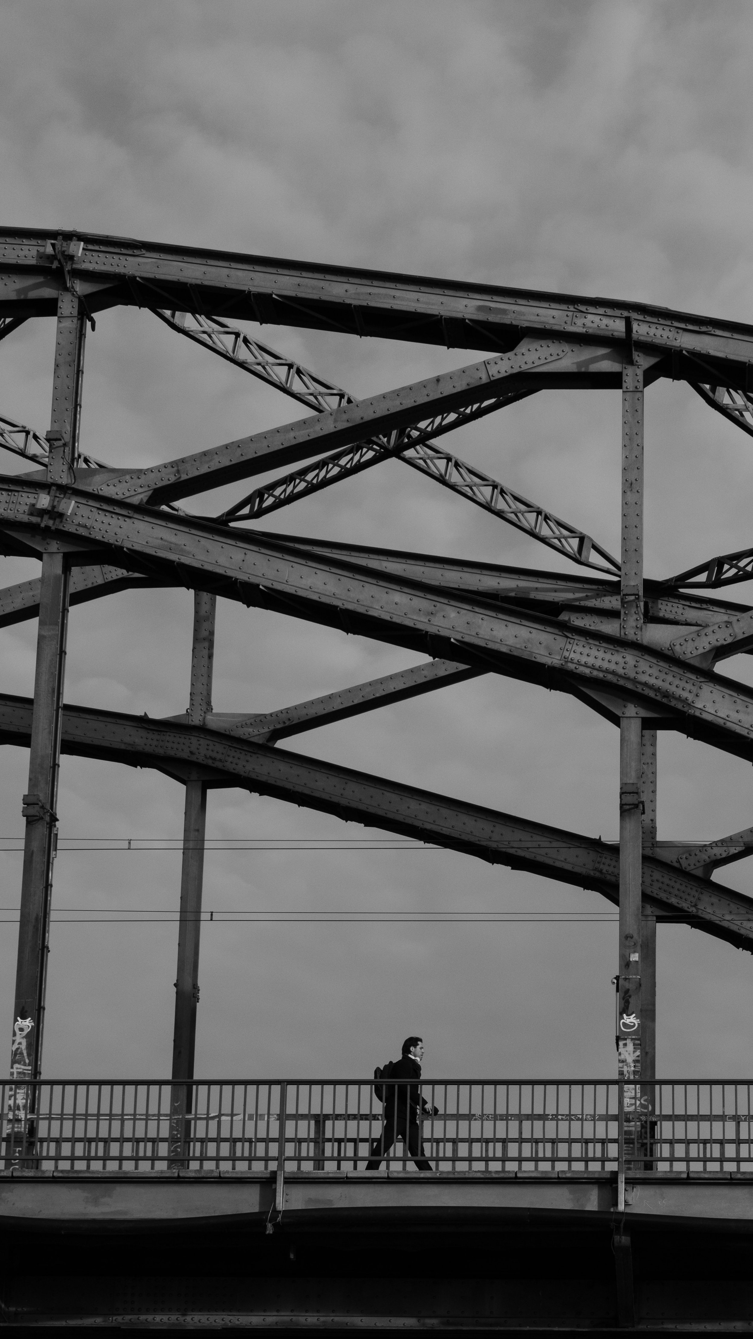 Schwarze und weiße Aufnahme einer Eisenbahnbrücke mit einem Mann, der auf der unteren Ebene geht, gegen den Hintergrund eines wolkigen Himmels.