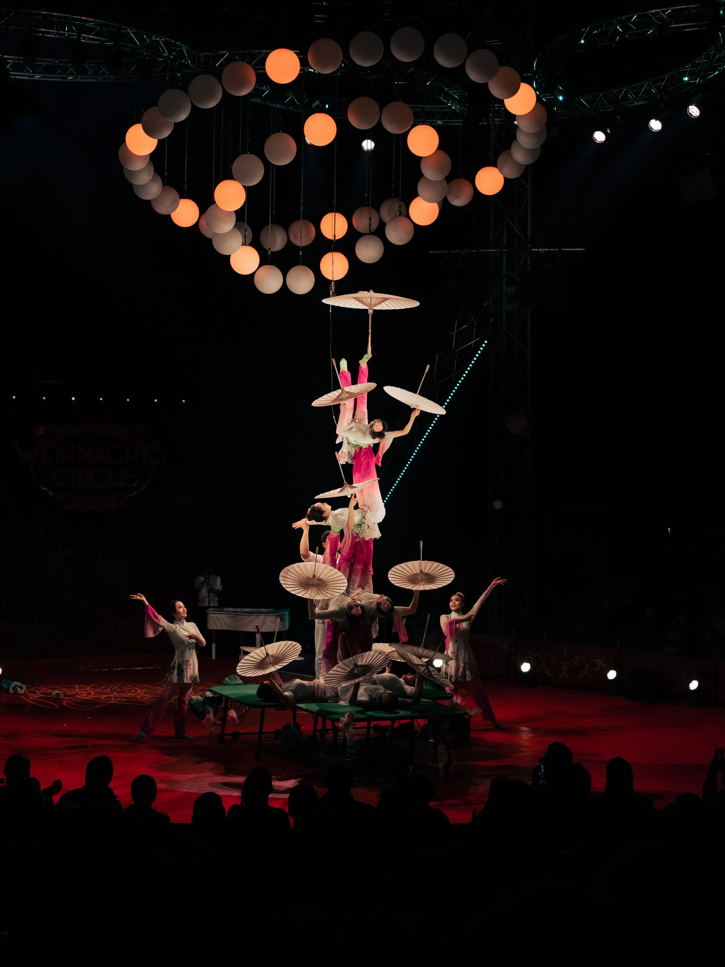 A circus performance with performers balancing and stacking umbrellas, illuminated by stage lights, with an audience watching. Event-Fotografie beim Mannheimer Weihnachtszirkus.