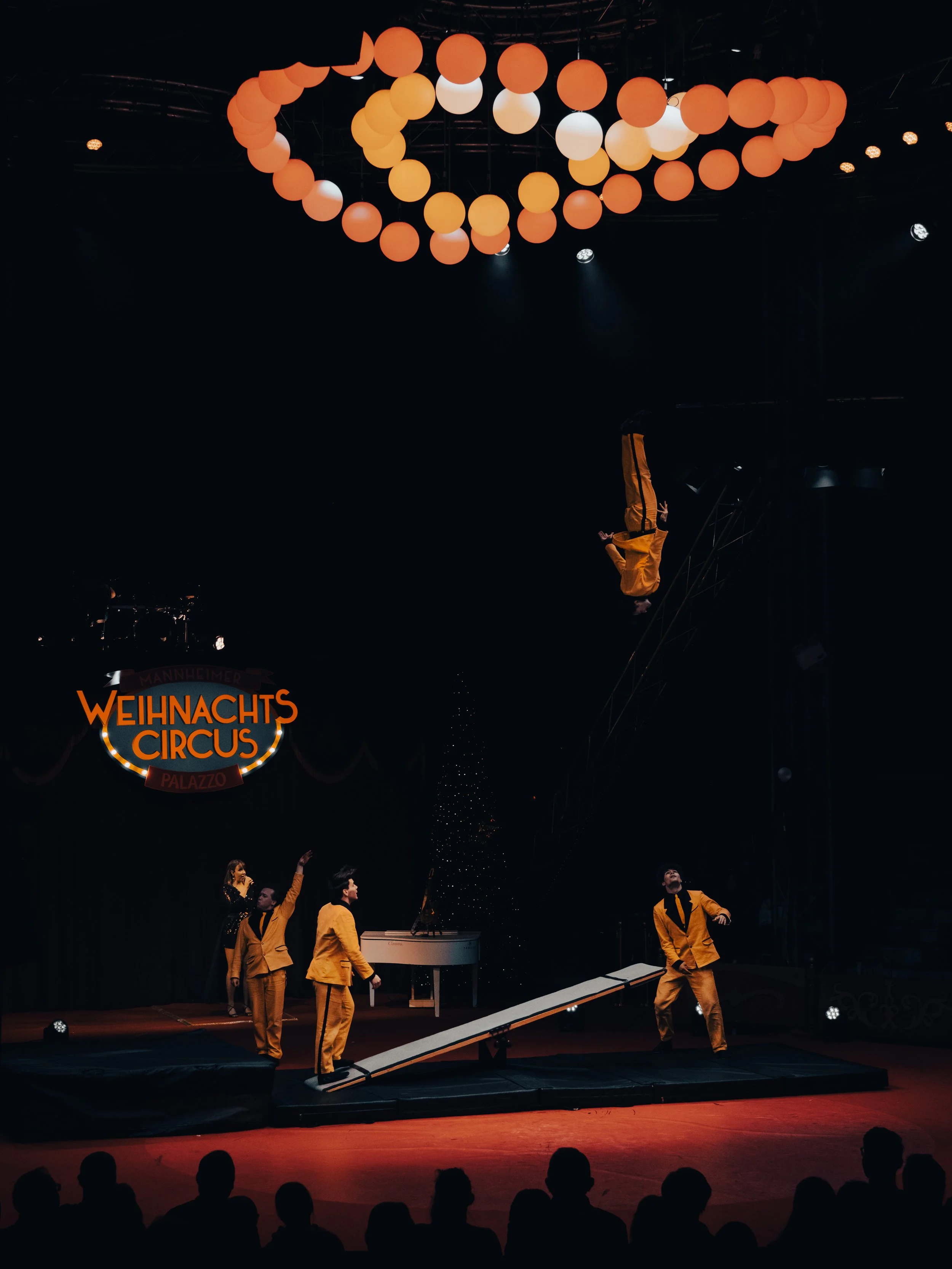 Zirkusperformance im Weihnachtscircus, mit Akrobaten in gelber Kleidung, die auf einer Bühne auftreten, während ein Akrobat auf einem Trampolin in der Luft ist, und ein Publikum im Dunkeln zuschaut.