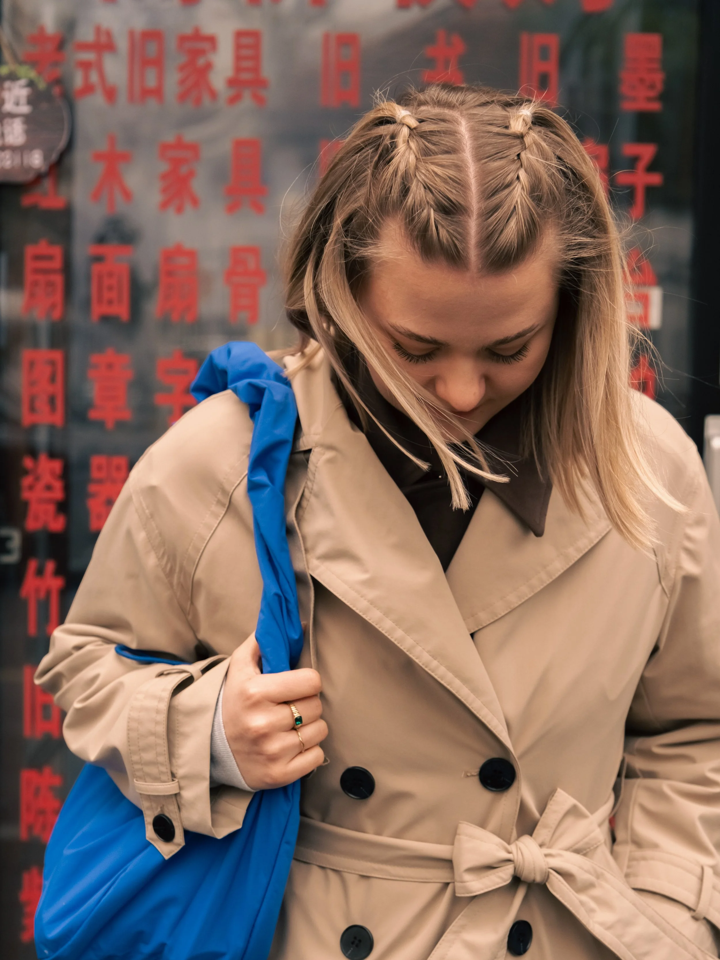 Eine junge Frau mit geflochtenen Haaren trägt einen beige Trenchcoat mit schwarzen Knöpfen und eine blaue Tasche auf der Schulter, vor einer Wand mit roten chinesischen Schriftzeichen.