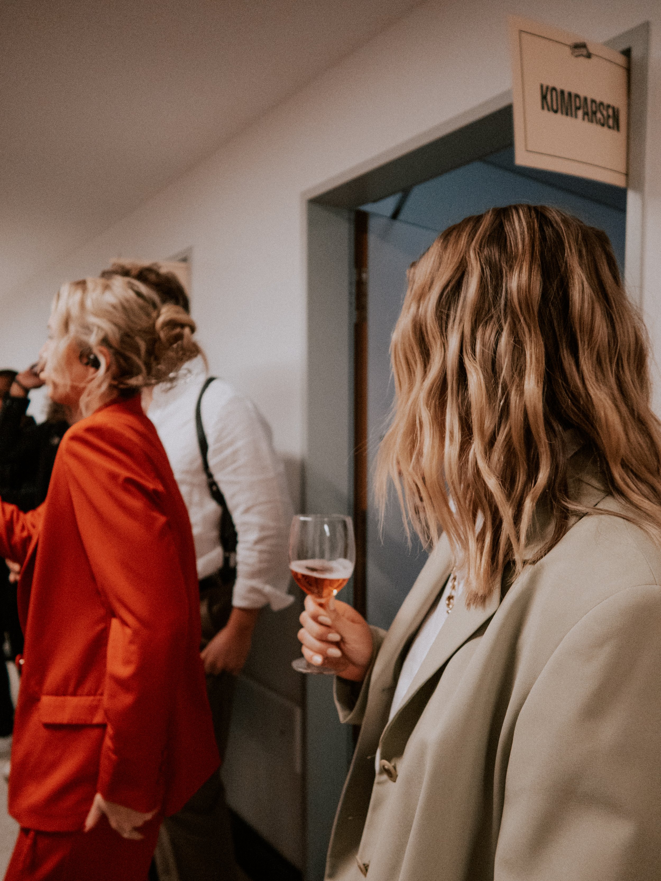 Menschen auf einer gesellschaftlichen Veranstaltung, eine Person hält ein Glas Roséwein, im Hintergrund ein Türschild mit der Aufschrift 'KOMPARSEN'.