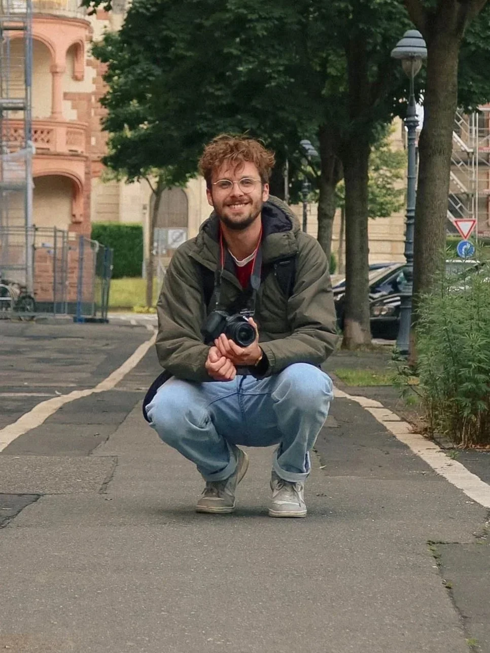 Ein lachender Mann mit Brille und lockigem Haar, der eine Kamera hält und in die Kamera kniet, auf einer städtischen Straße mit Bäumen und Gebäuden im Hintergrund.