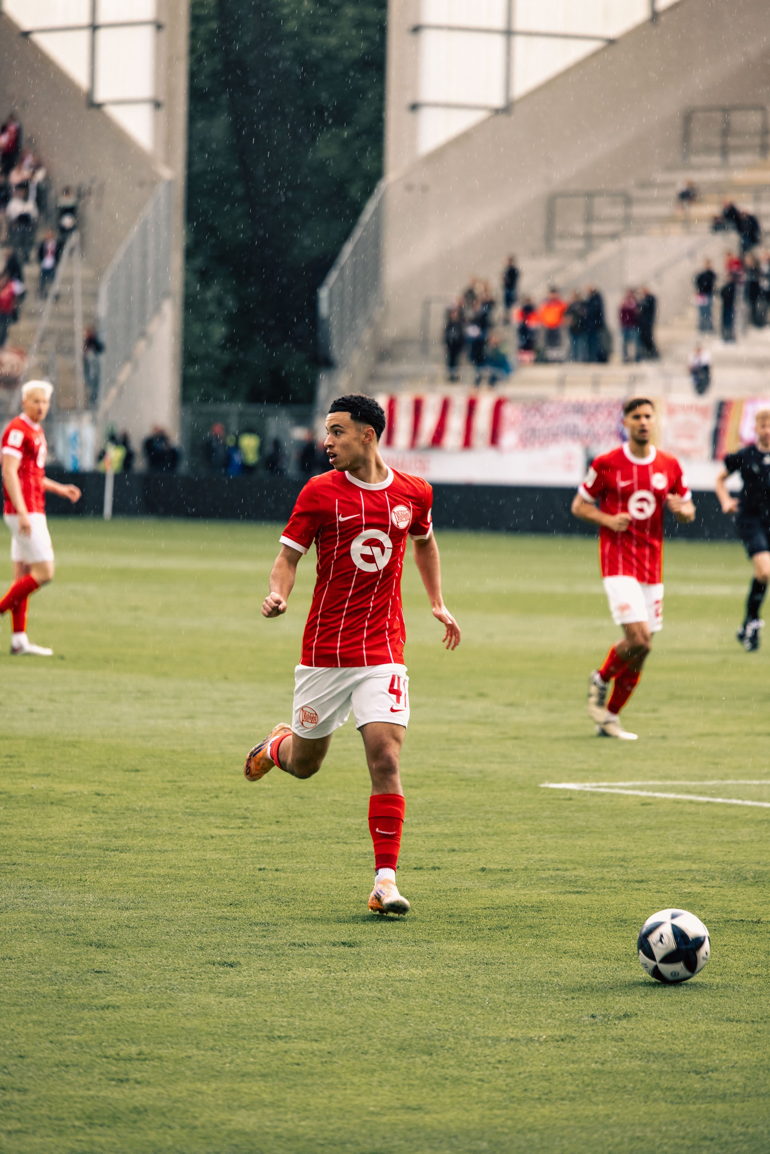 Fußballspieler der Kicker Offenbach in rotem Trikot läuft auf dem Spielfeld, während im Hintergrund andere Spieler und Zuschauer im Stadion zu sehen sind. Sportfotografie bei den Kickers Offenbach.