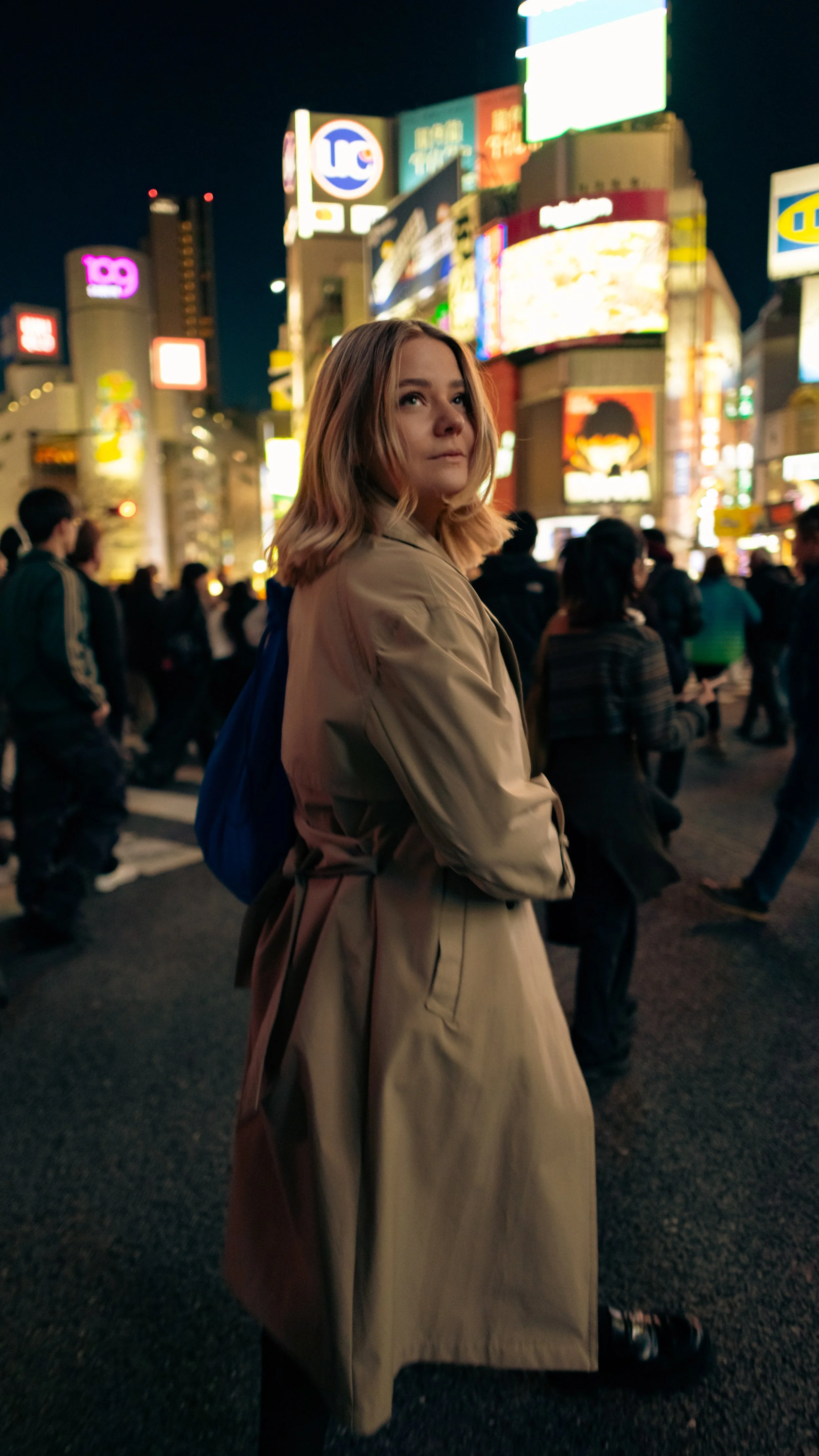 Junge Frau mit blonden Haaren in einem hellbraunen Trenchcoat steht in einer belebten, nächtlichen Stadt auf der Shibuya-Kreuzung  mit leuchtenden Reklametafeln.
