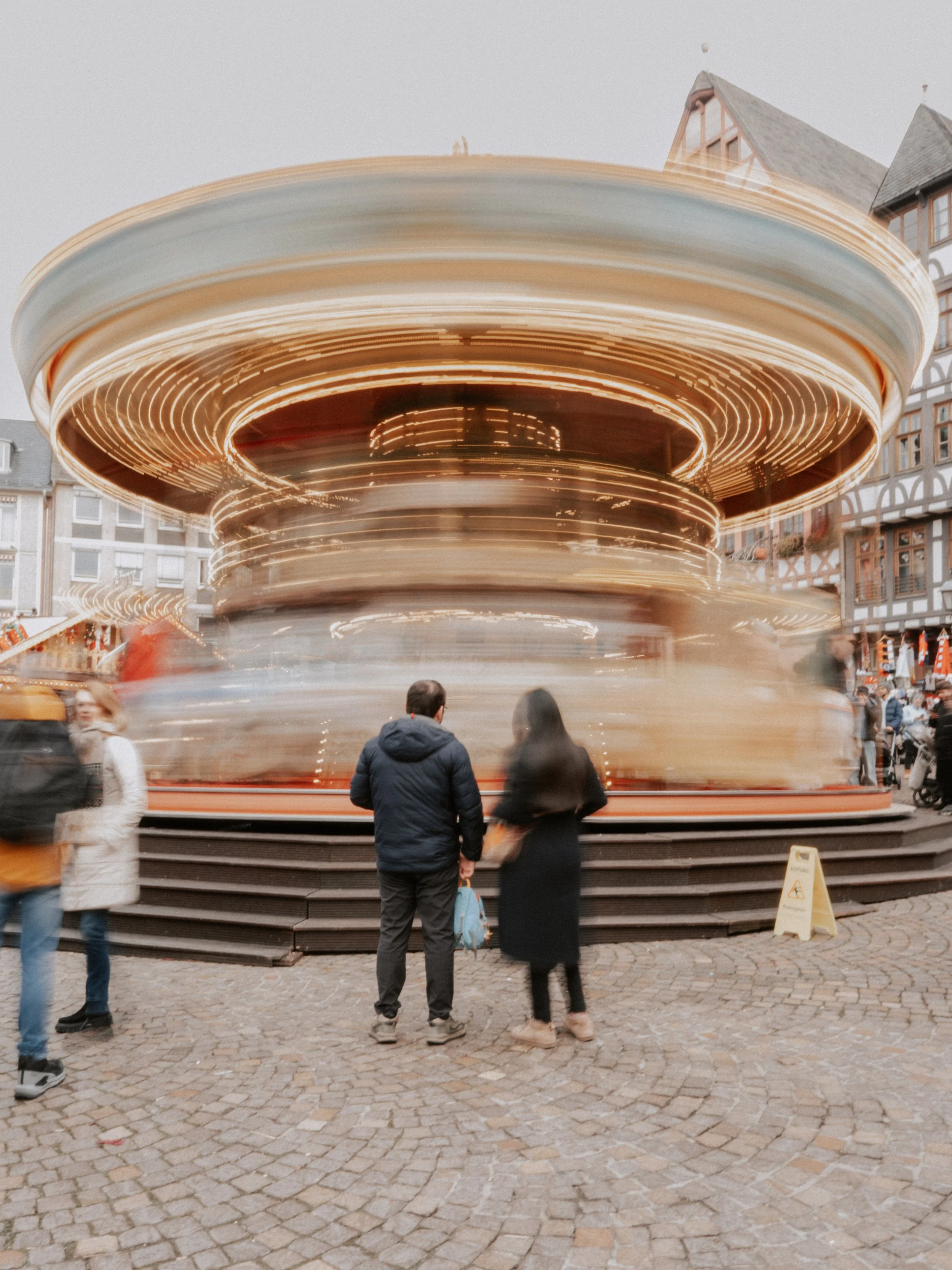 Menschen stehen vor einer beleuchteten, drehenden Karussell-Fahranlage auf einem Rathausmarkt. Das Bild ist leicht verschwommen durch Langzeitbelichtung bzw. Slow Shutter.