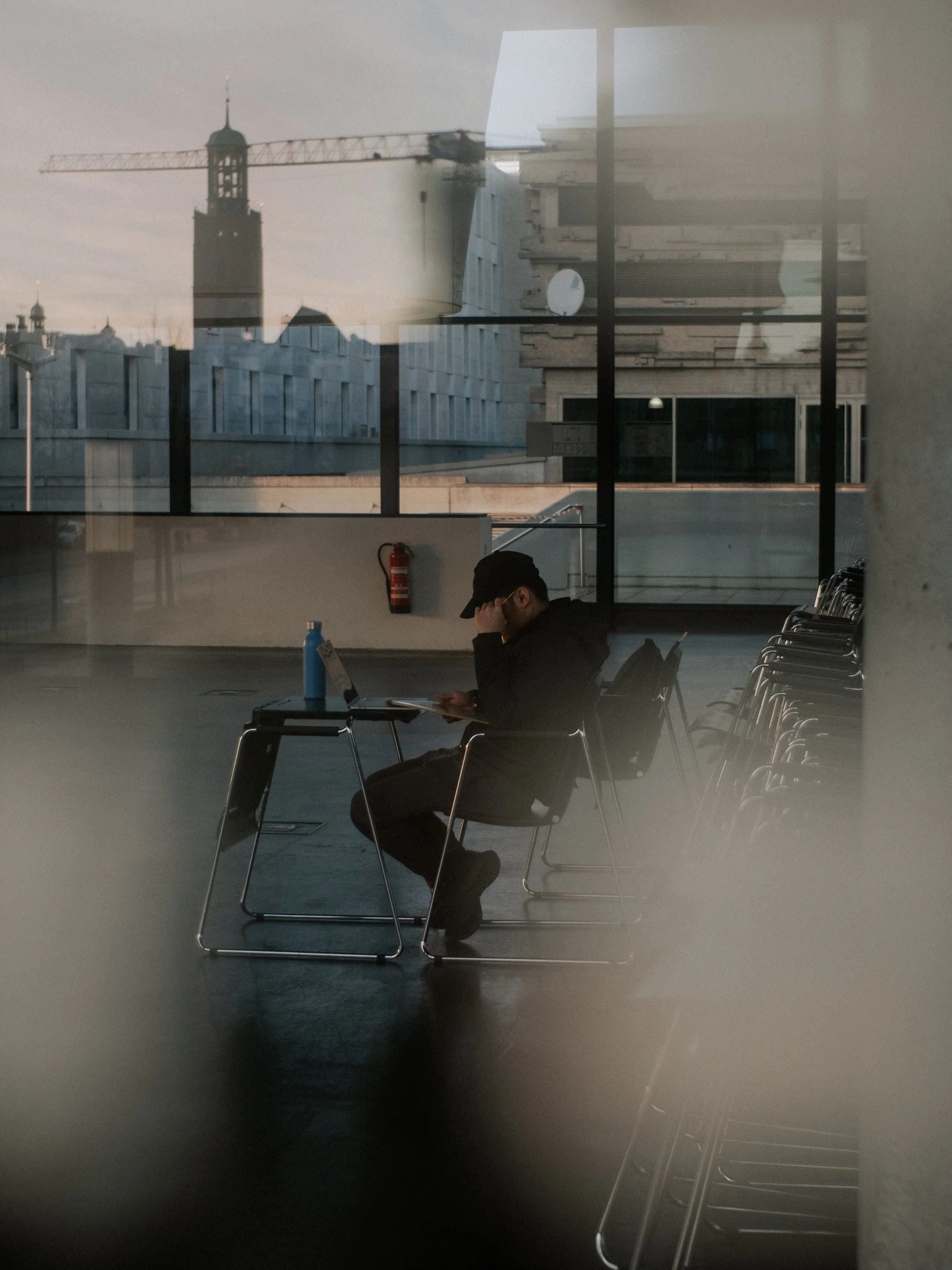 Im Inneren eines Gebäudes, mit einer Person, die auf einem Stuhl sitzt und auf ein Mobiltelefon schaut, während andere leere Stühle neben ihr stehen. Durch das Fenster ist eine städtische Skyline mit einem Baukran und einem Turm zu sehen.