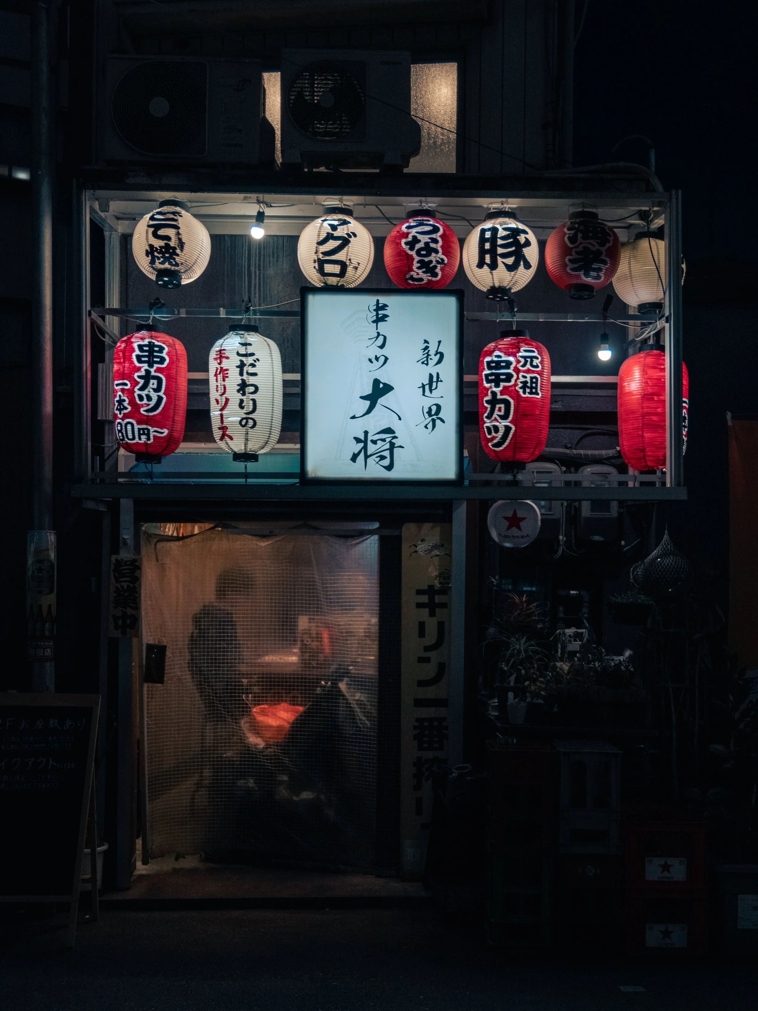 Ein Nachtbild einer kleinen Essensbude mit leuchtenden japanischen Laternen, die Namen und Speisen in japanischer Schrift tragen, darunter eine große weiße Tafel mit japanischen Schriftzeichen.