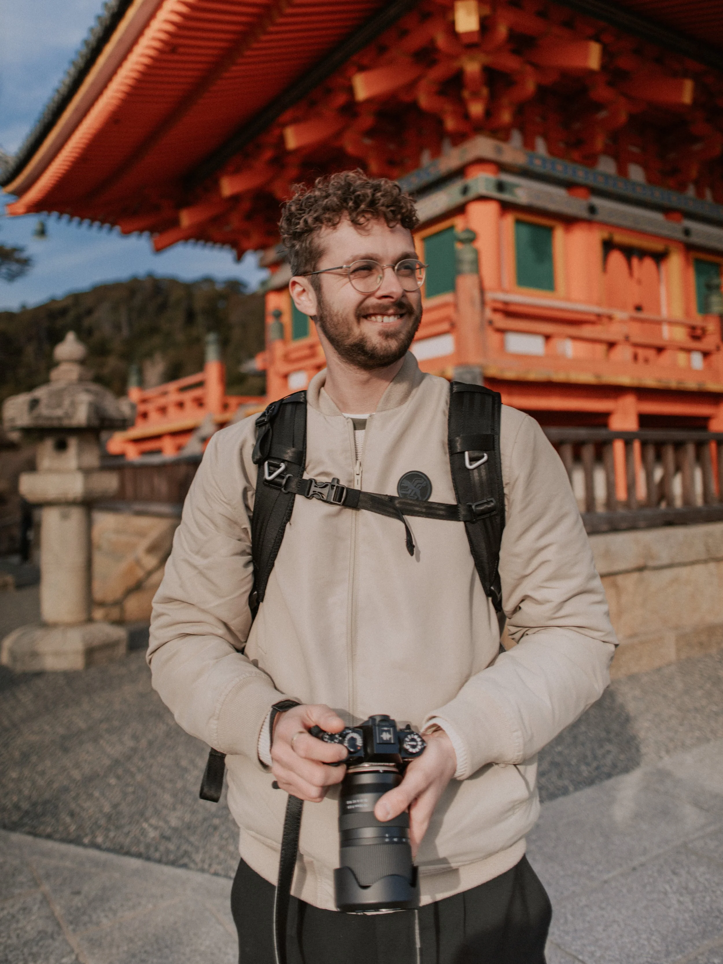 Junger Mann mit Brille und Kamera in den Händen, stehend vor einer traditionellen japanischen Tempelanlage bei Sonnenuntergang.