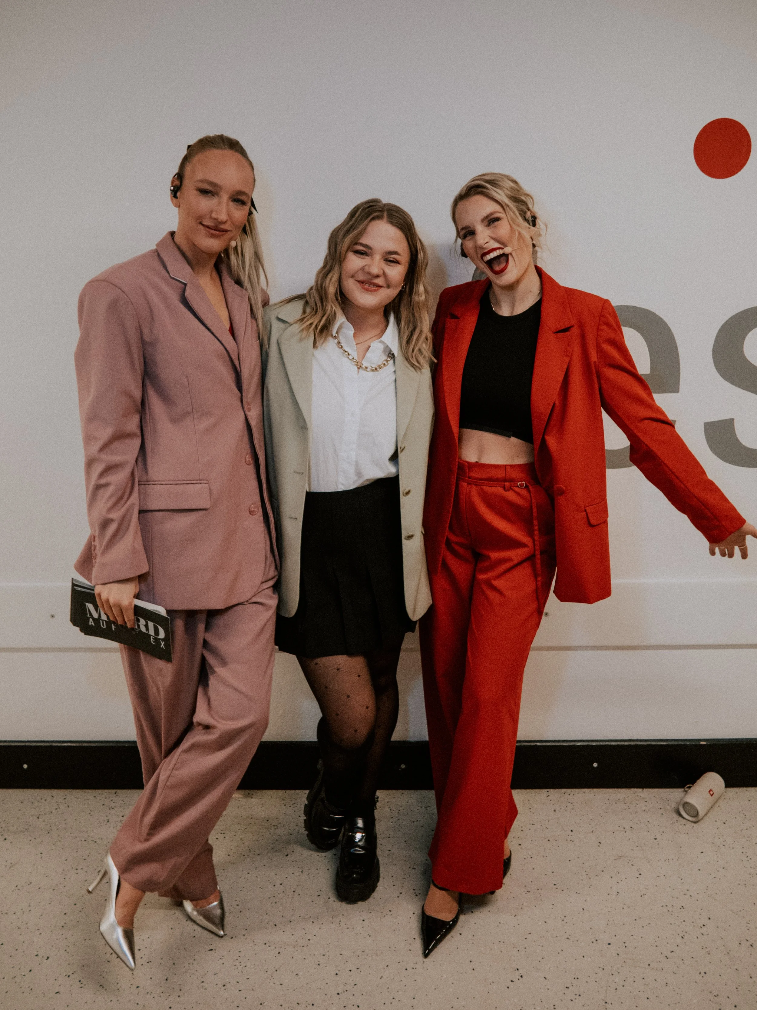 Drei Frauen in eleganten Outfits beim Posieren und Lachen vor einer weißen Wand.