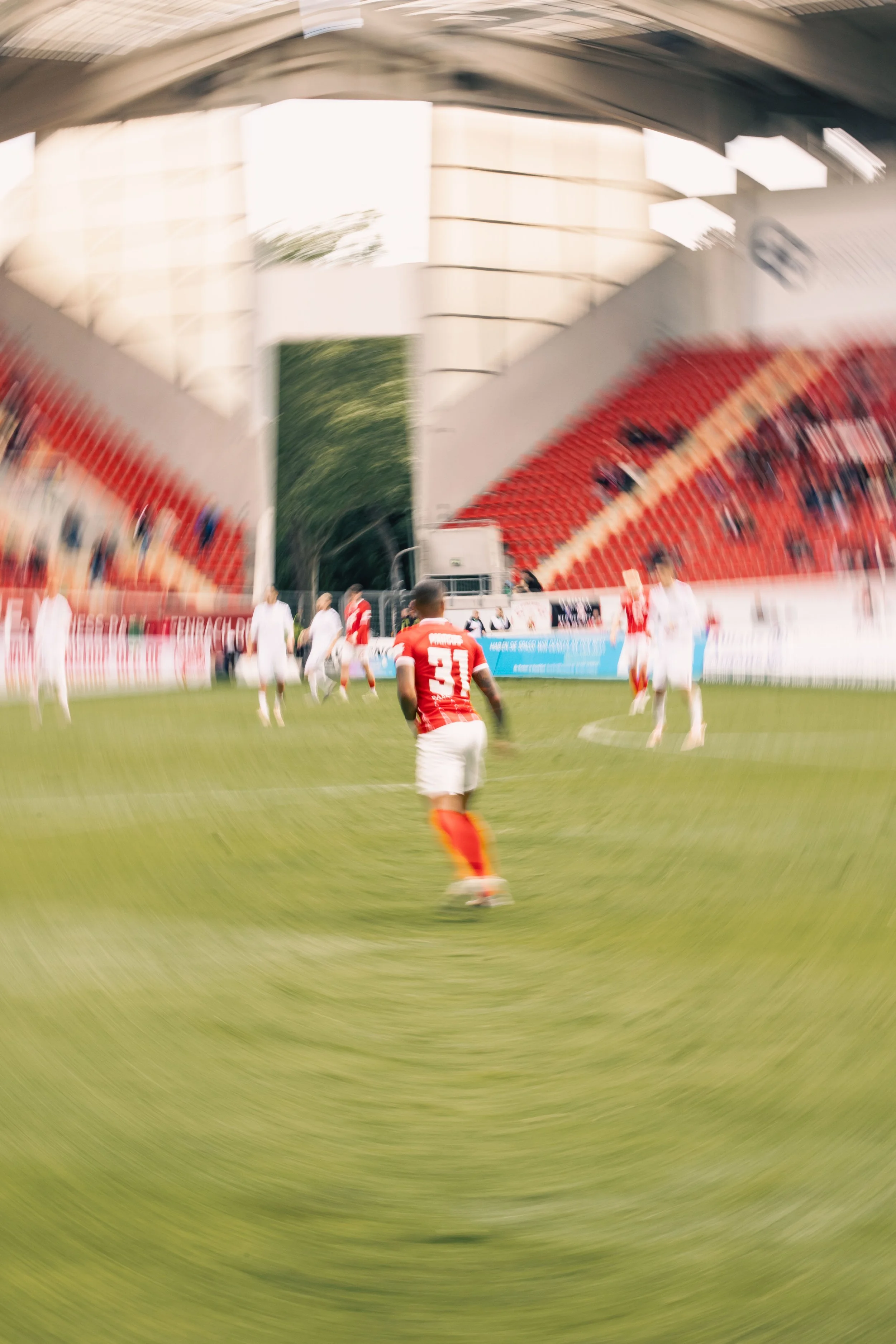 Fußballspieler in roter Trikotnummer 33 auf einem grünen Spielfeld, im Hintergrund rot-weiße Sitzreihen im Stadion, der Bildausschnitt ist unscharf durch Langzeitbelichtung.