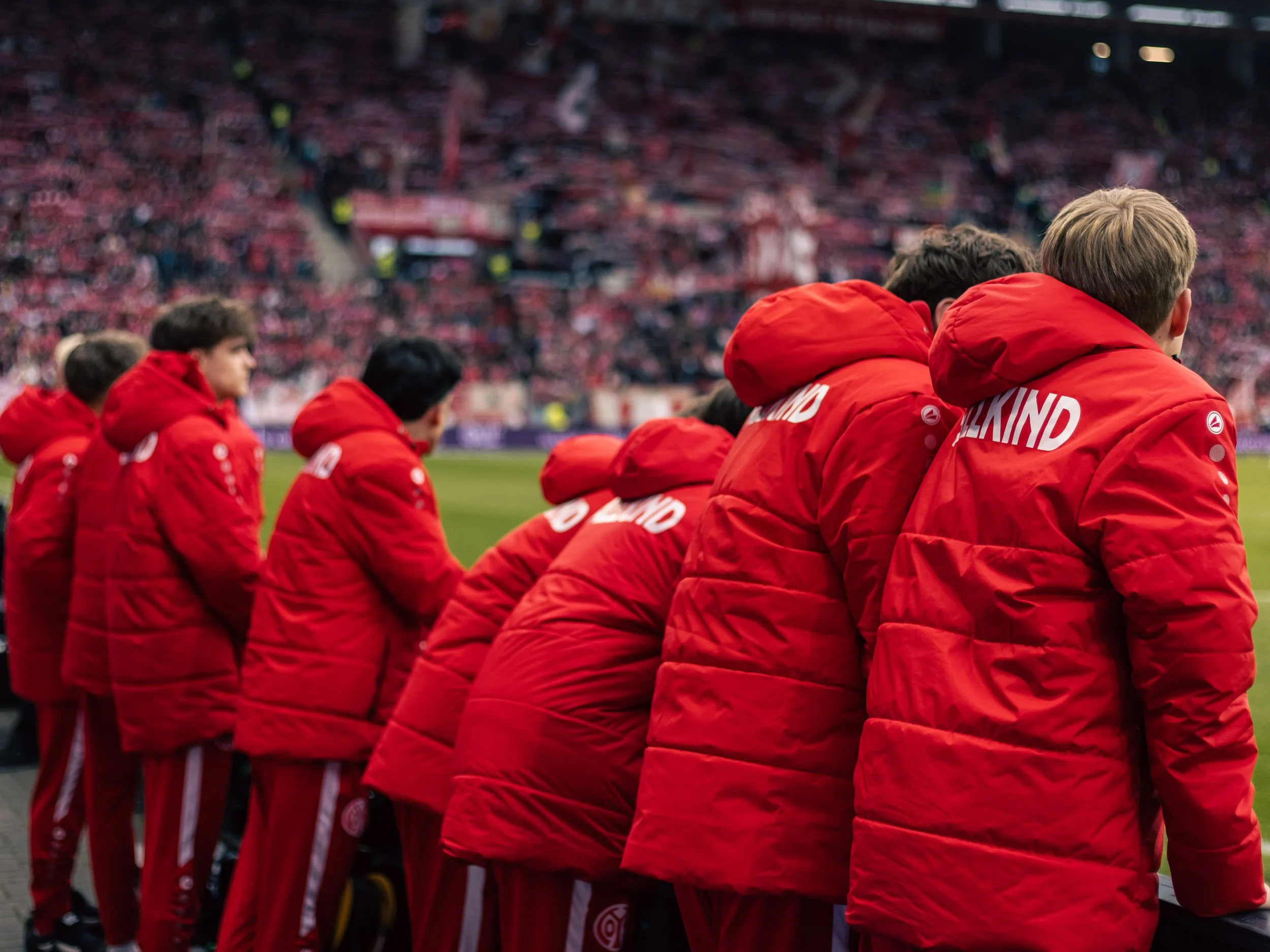 Balljungen beim 1. FSV Mainz 05 in roten Jacken, die nebeneinander am Spielfeldrand stehen, während eines Spiels, mit einem vollen Stadion im Hintergrund. Sportfotografie beim 1. FSV Mainz 05 in der Bundesliga.