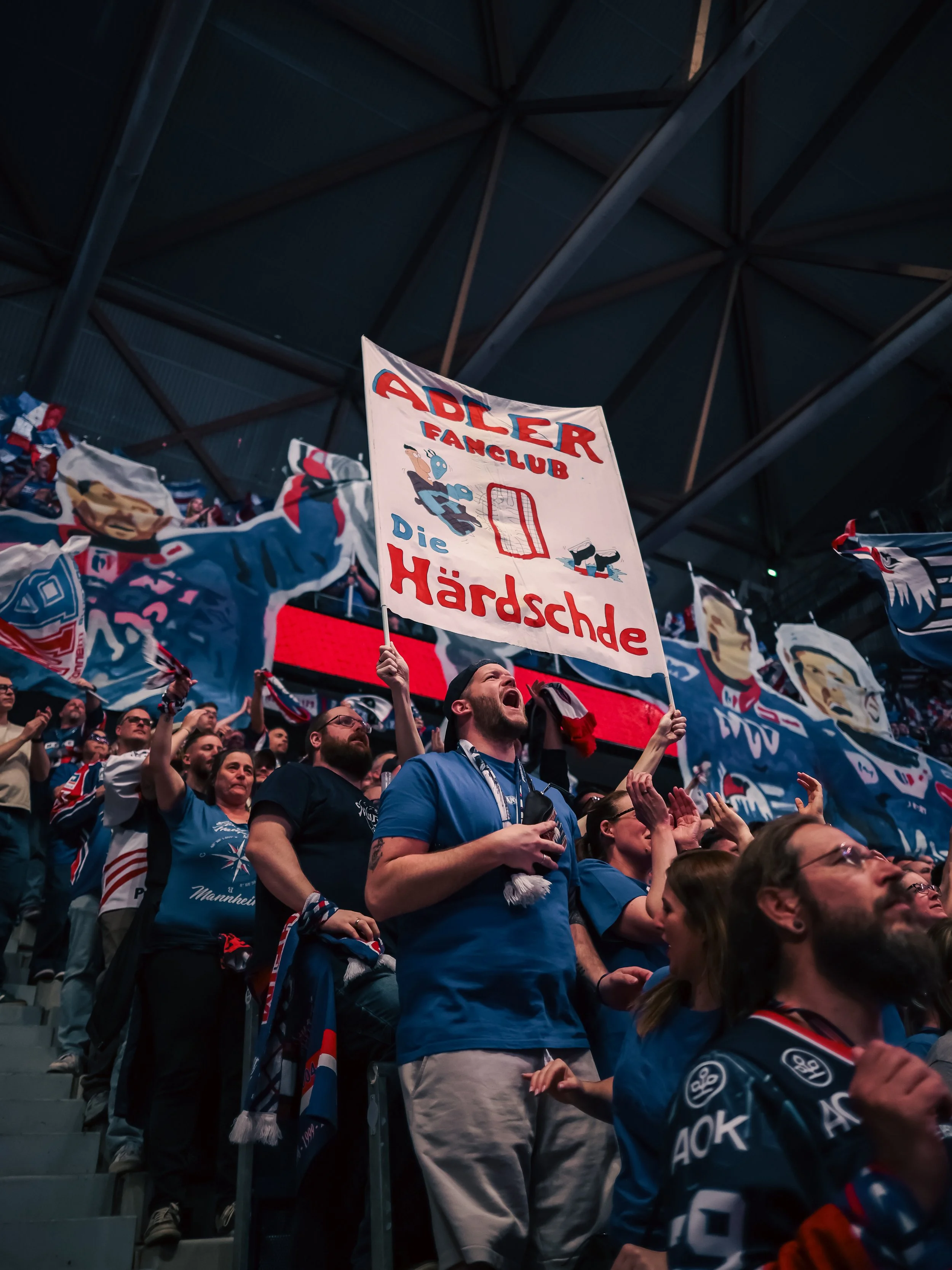 Fans feiern in einer Halle, einige halten Schilder und Banner, eines mit Aufschrift 'Adler Fanclub Die Härteschde', während sie jubeln und anfeuern, gekleidet in Fan- und Mannschaftskleidung.