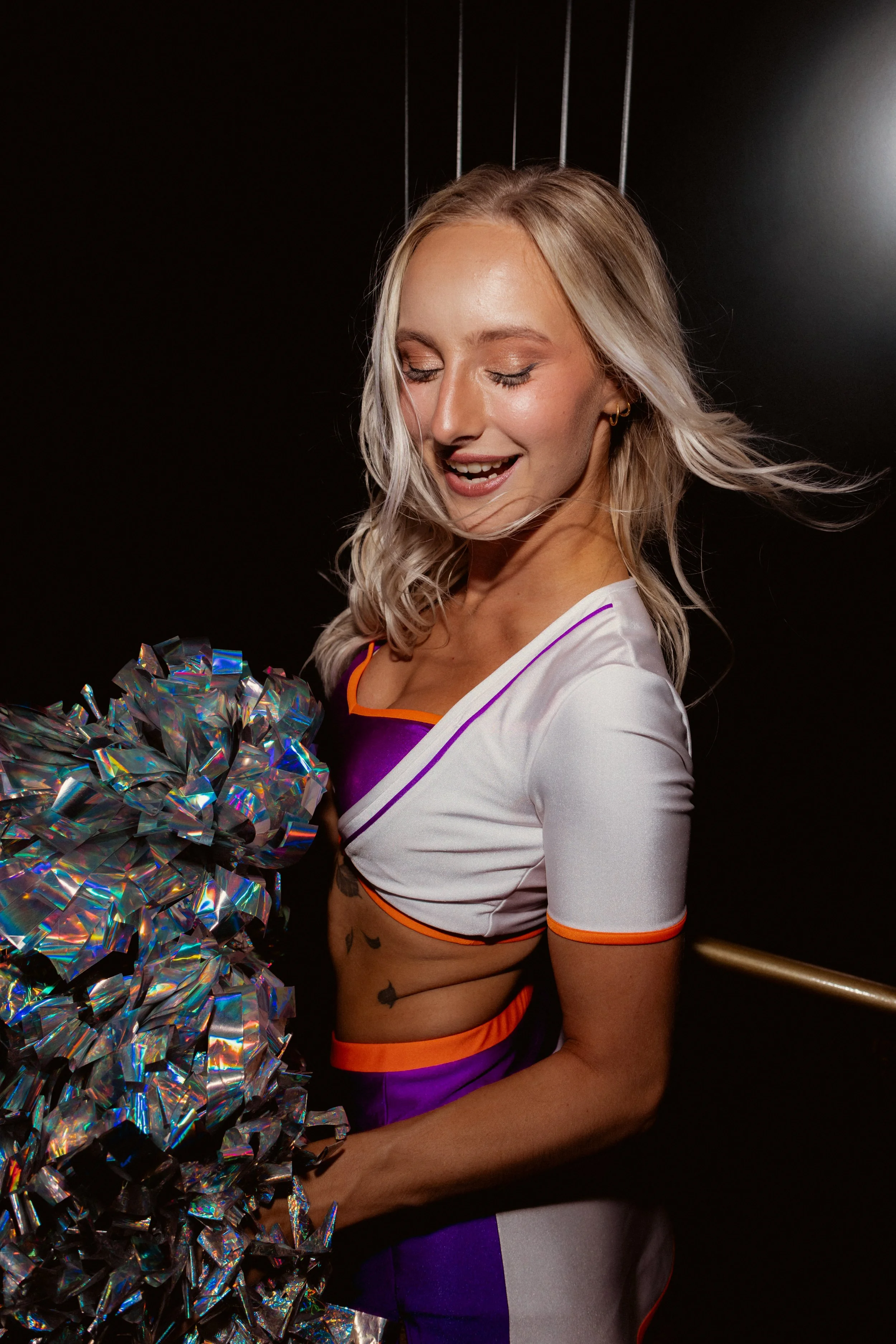 Lächelnde Cheerleaderin mit blonden, lockigen Haaren, in einem weißen Outfit mit lila und orange Akzenten, hält silberne pompons, steht vor einem schwarzen Hintergrund