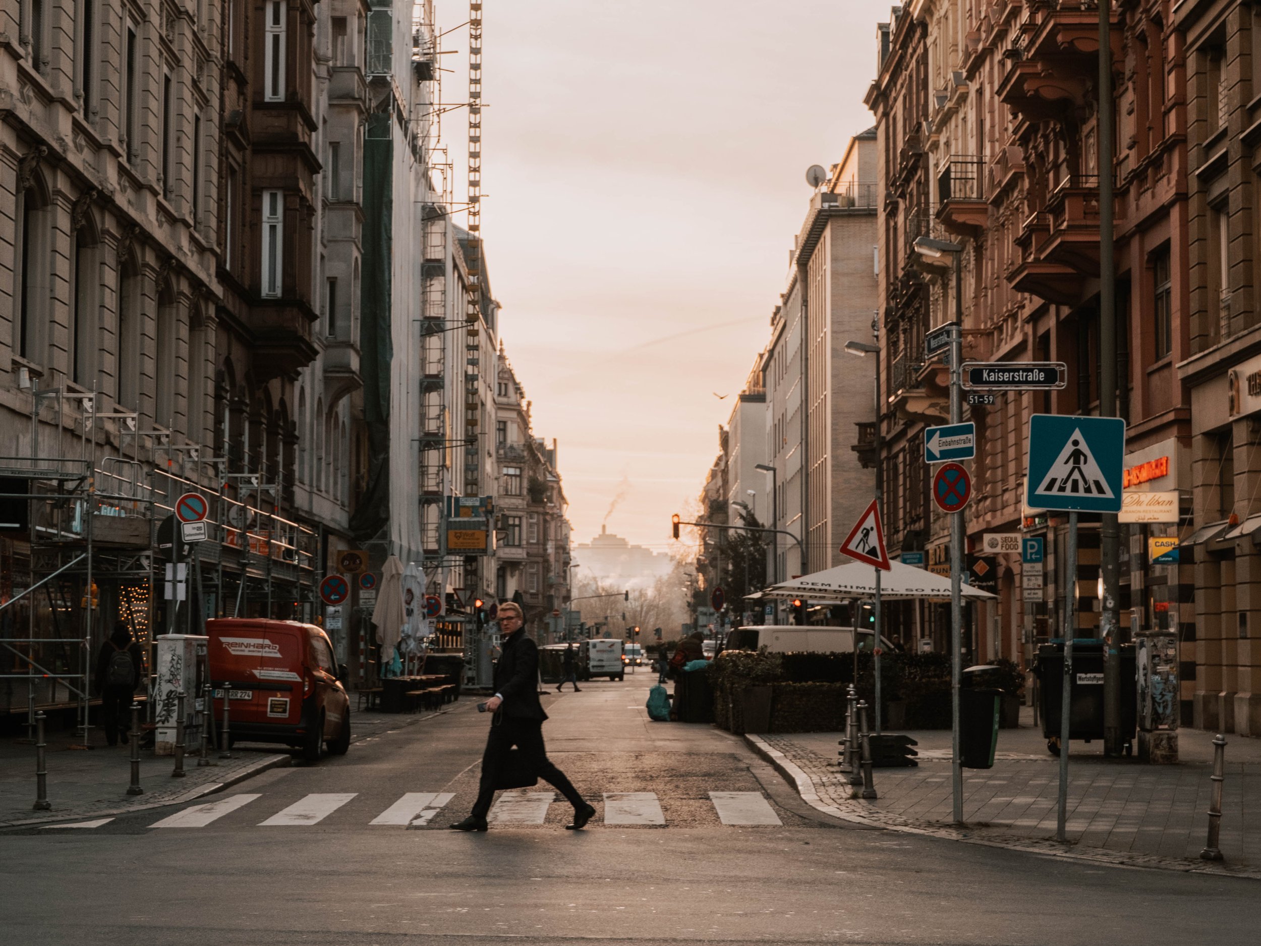 Straßenkreuzung in einer Stadt bei Sonnenuntergang mit mehreren mehrstöckigen Gebäuden, Fußgängerweg, Ampeln, Verkehrsschildern und wenigen Menschen auf der Straße in Frankfurt am Main.