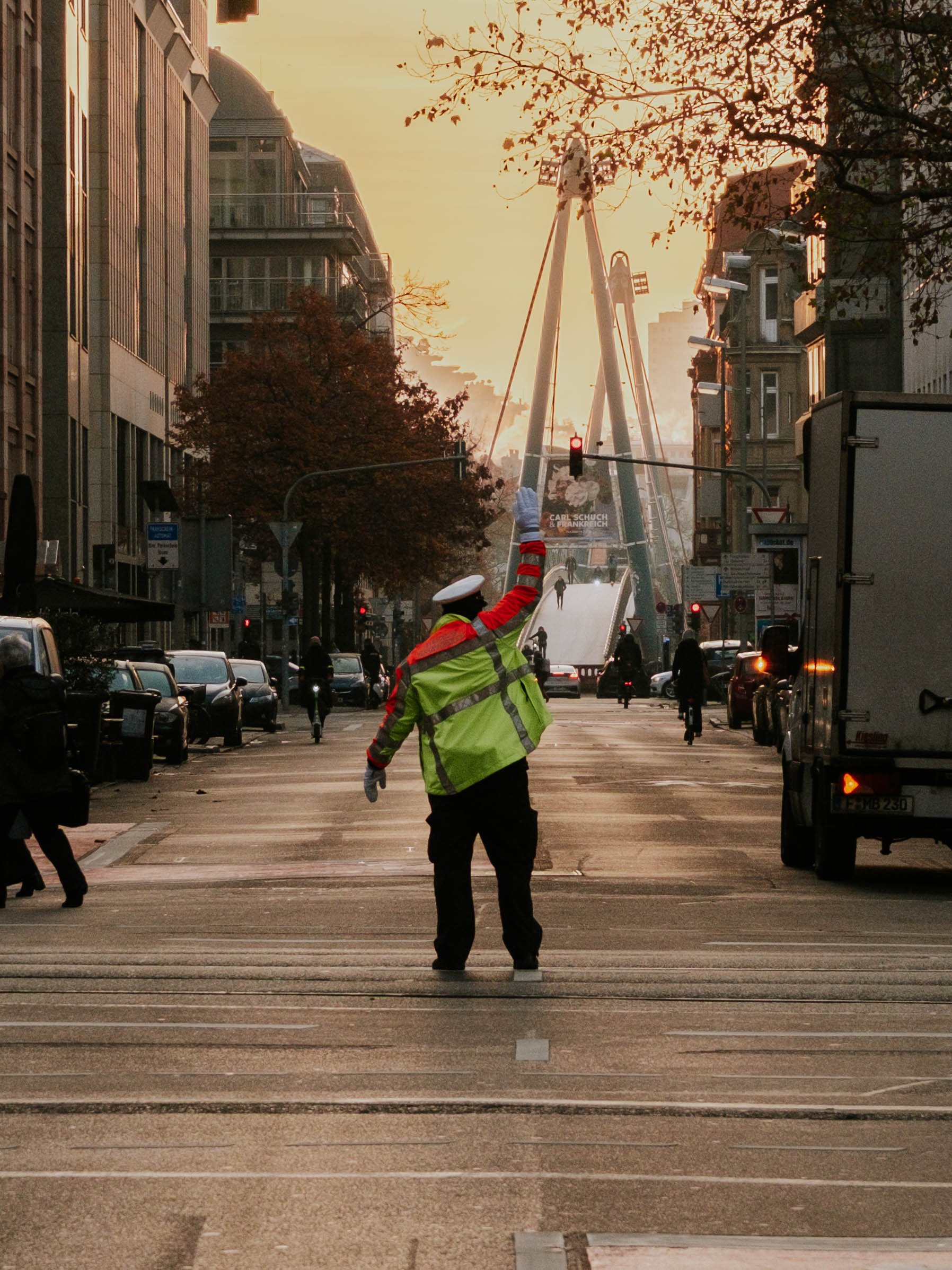 Straßenszene mit einem Polizisten der den Verkehr regelt, im Hintergrund Sonnenuntergangslicht, Bäume und Gebäude auf beiden Seiten der Straße. Streetphotography in Frankfurt.