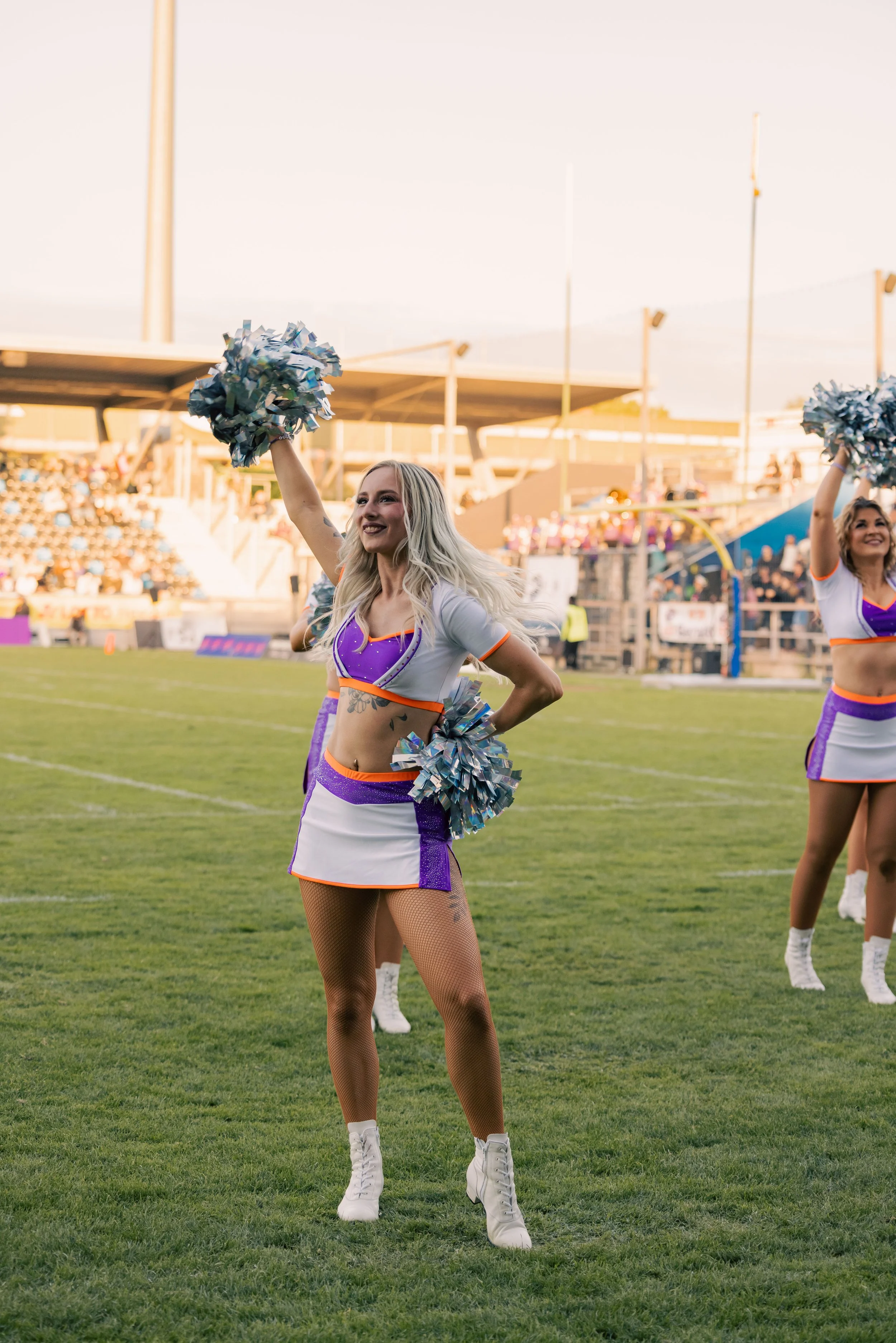 Cheerleader auf einem Sportfeld bei Sonnenuntergang, trägt eine lila-weiße Uniform mit Orange-Akzenten, hält silberne Pompoms, lächelt, trägt Netzstrümpfe und weiße Turnschuhe, im Hintergrund ist die Tribüne mit Zuschauern zum Teil sichtbar.