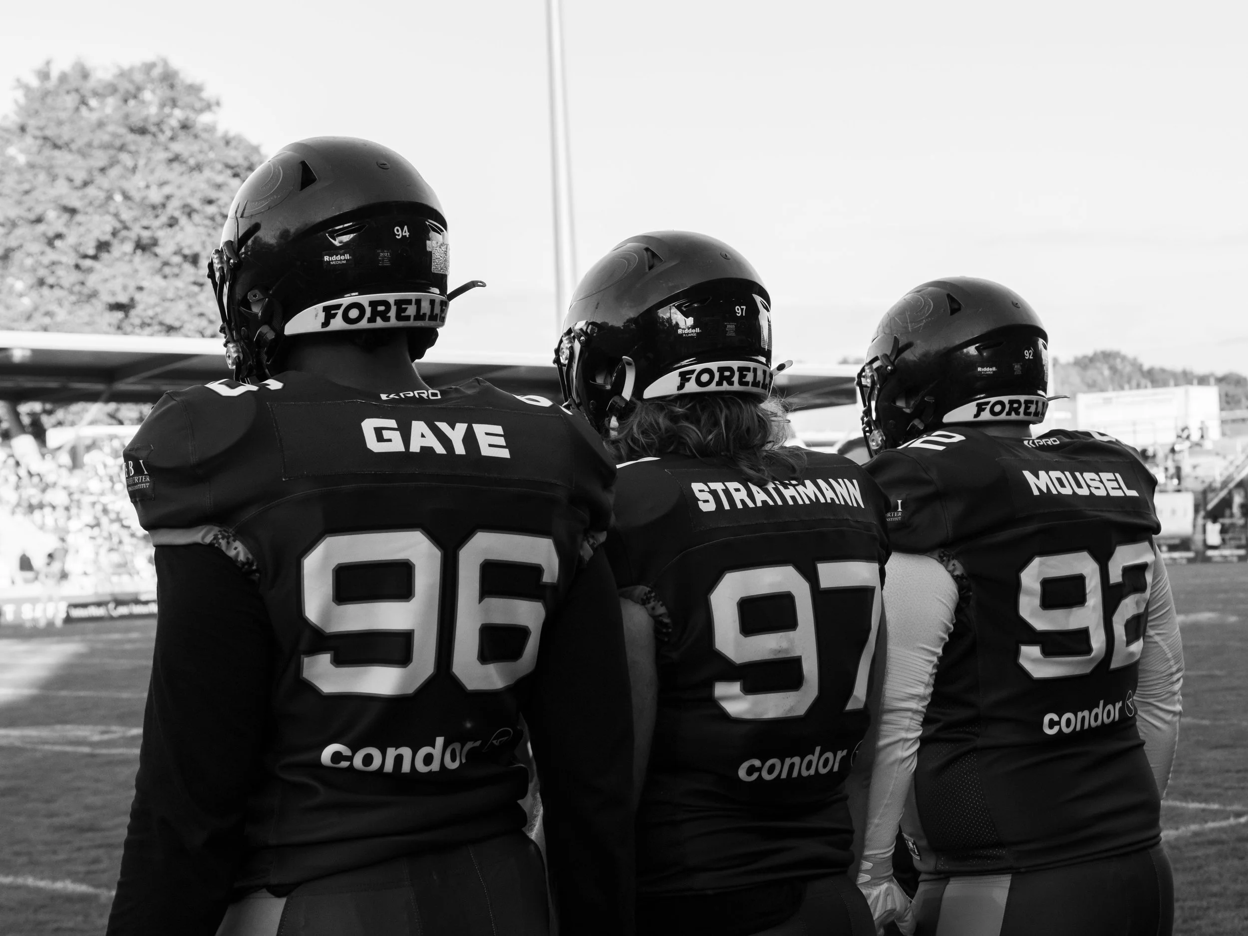 American-Football-Spieler stehen nebeneinander auf dem Spielfeld, nach hinten blickend, und schauen in Richtung der Spielfeldmitte. Sie tragen Helme und Trikots mit den Namen Gaye, Strachmann, Mousel, und eine Nummer, sowie den Sponsor-Text "condor".