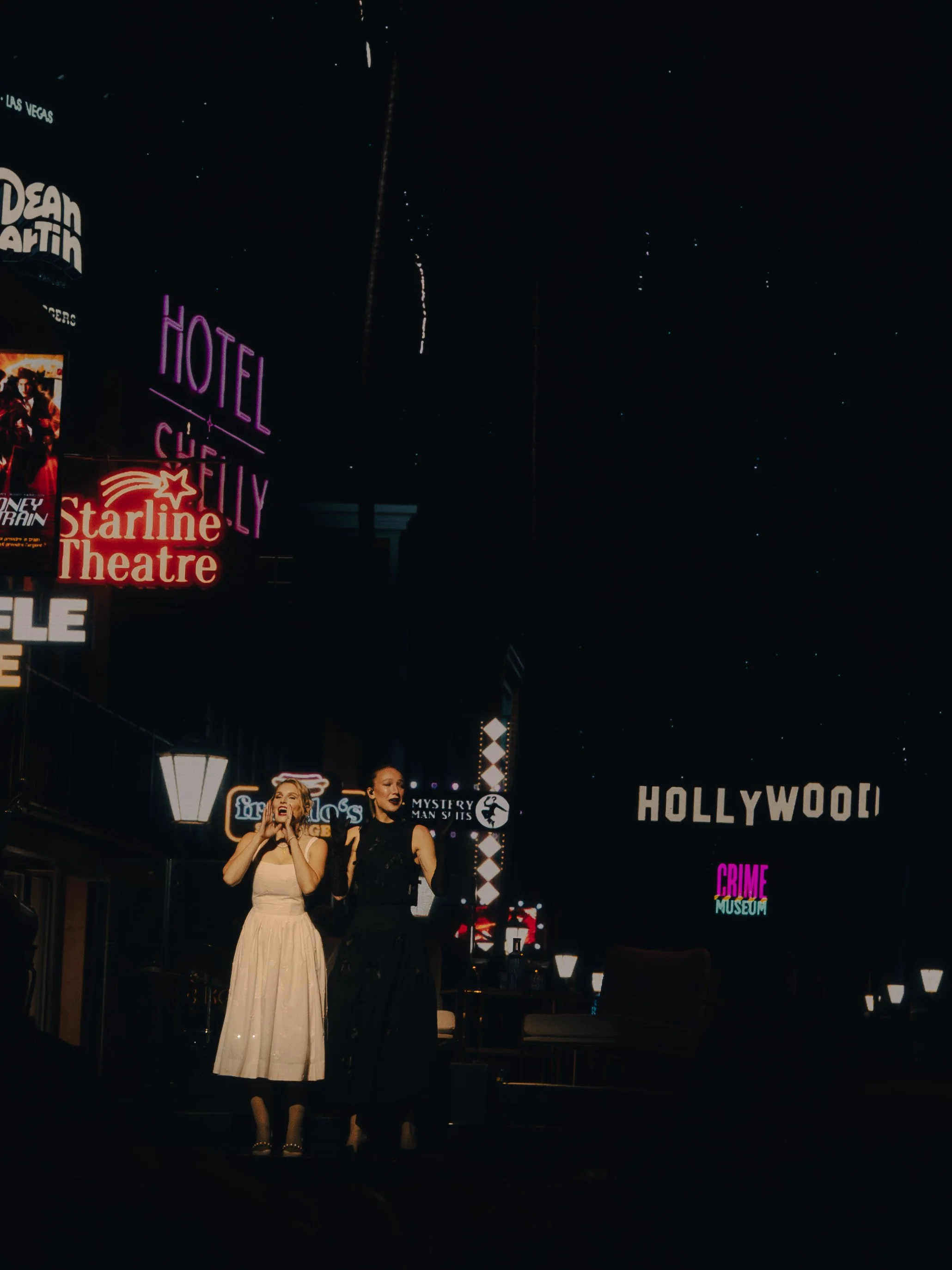 Zwei Frauen stehen auf einer Straßentreppe bei Nacht, im Hintergrund Neon-Reklameschilder für Hotels, Theatern und das Hollywood-Zeichen, Sterne am Himmel.