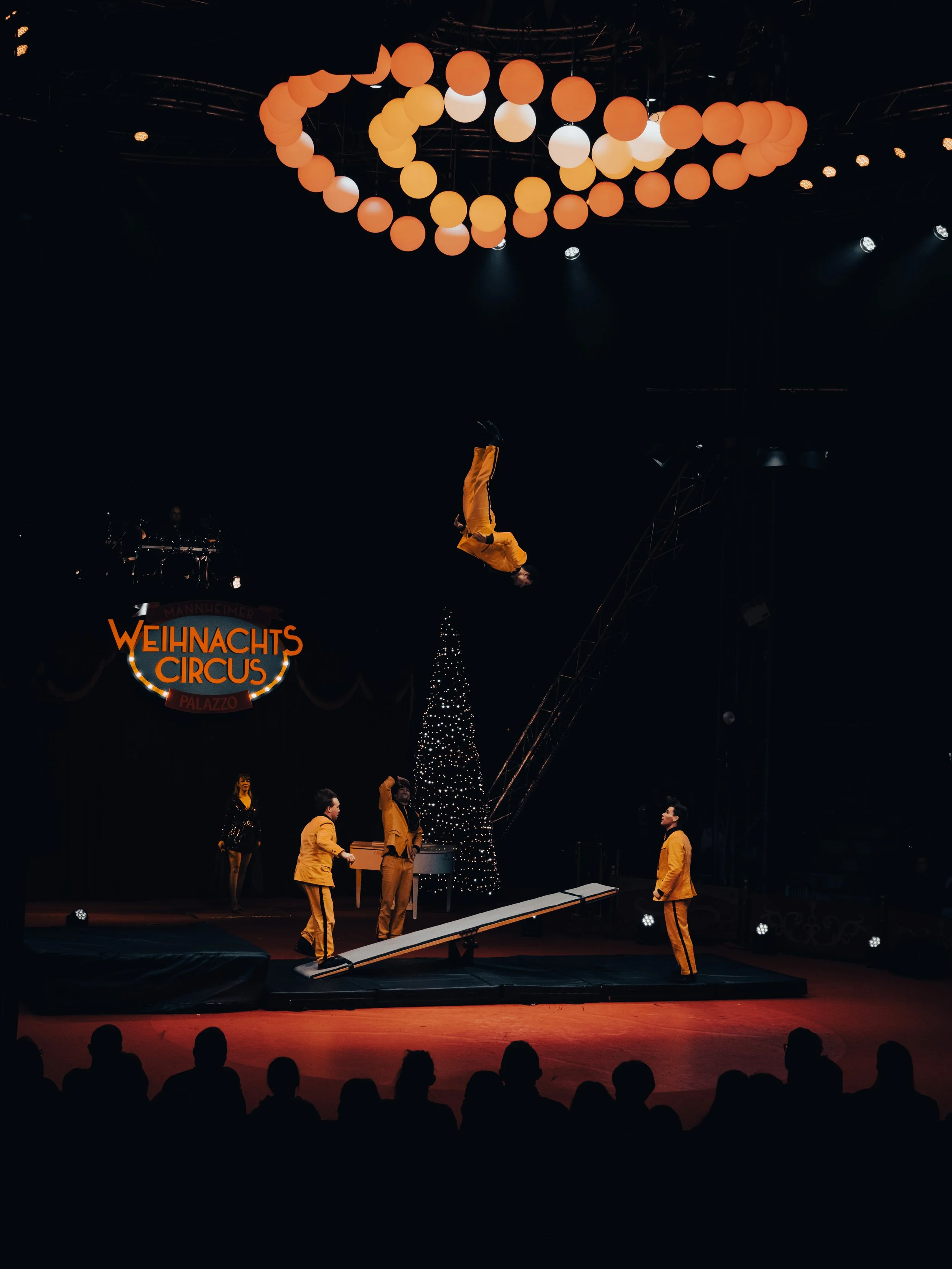 Zirkusaufführung mit einem Artist in gelber Kleidung, der in der Luft schwebt, während das Publikum im dunklen Saal zuschaut. Im Hintergrund sind Lichter, ein Weihnachtsbaum und eine Zirkusreklame zu sehen.