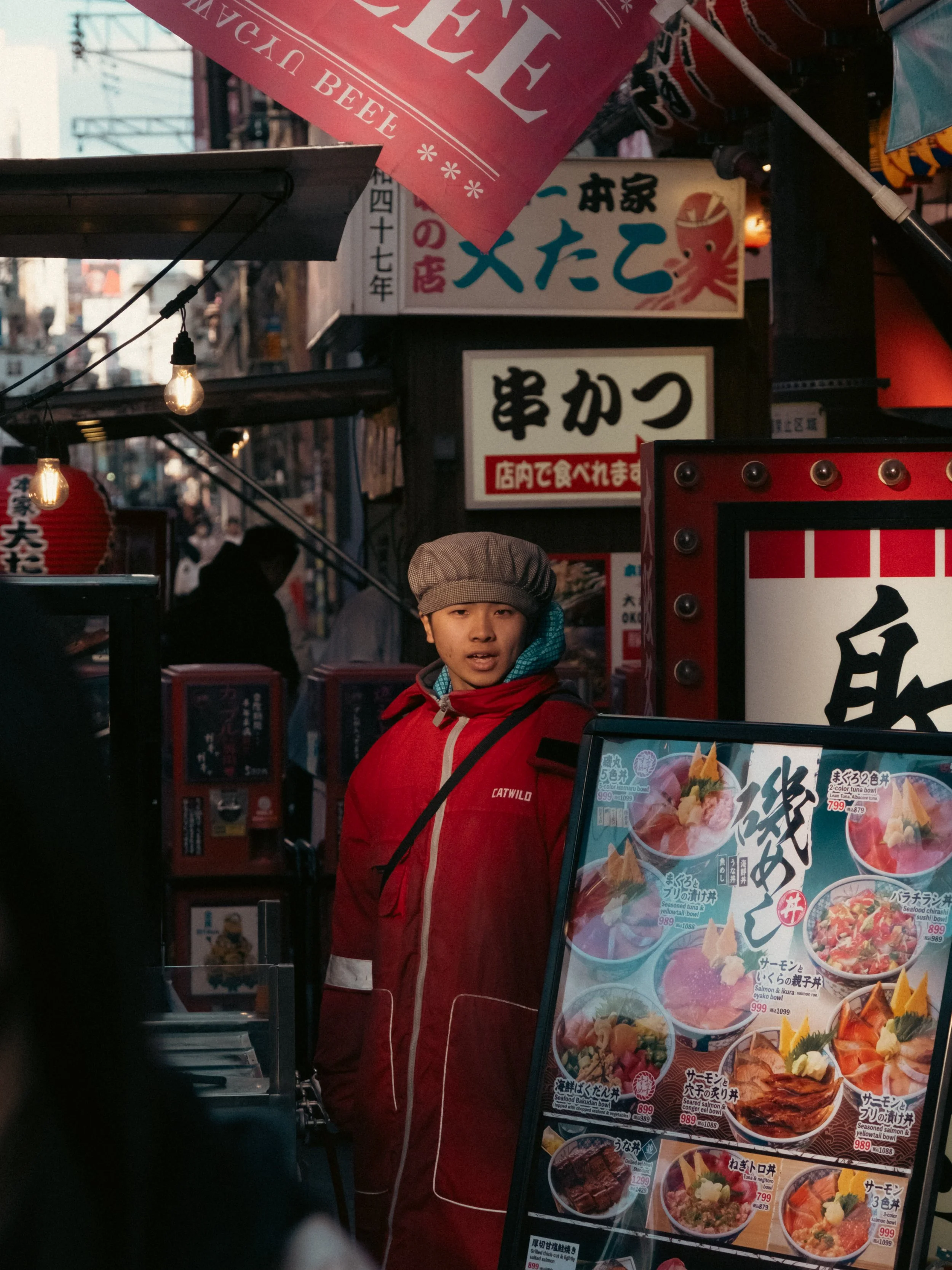 Straße in Osaka, Japan, eine Person trägt eine rot-blau Jacke und eine braune Mütze, im Hintergrund sind Lichter und Stadtgebäude zu sehen. Street-Fotografie in Japan.
